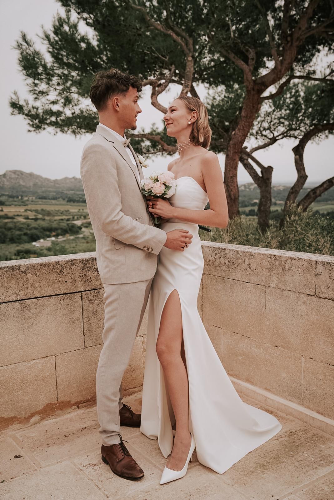 Smiling couple in wedding attire stands on stone terrace with scenic countryside and pine tree backdrop.