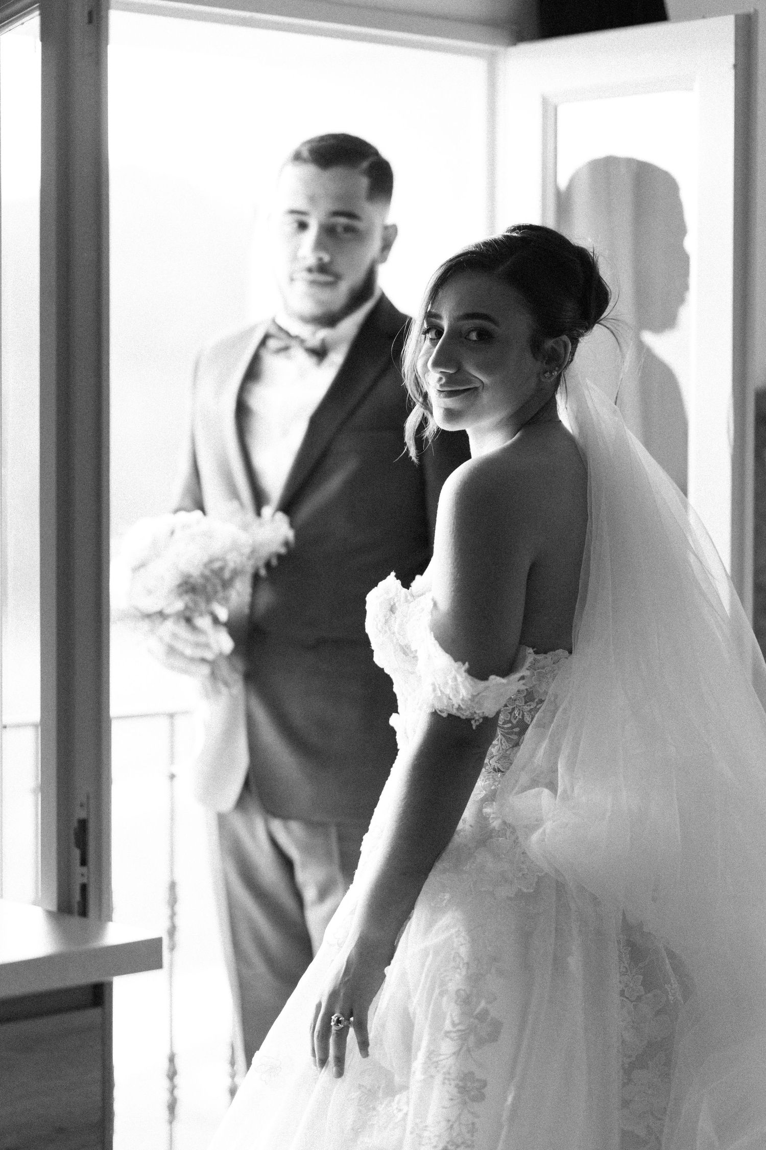 Black and white portrait of smiling bride in veil with groom holding bouquet by bright window.
