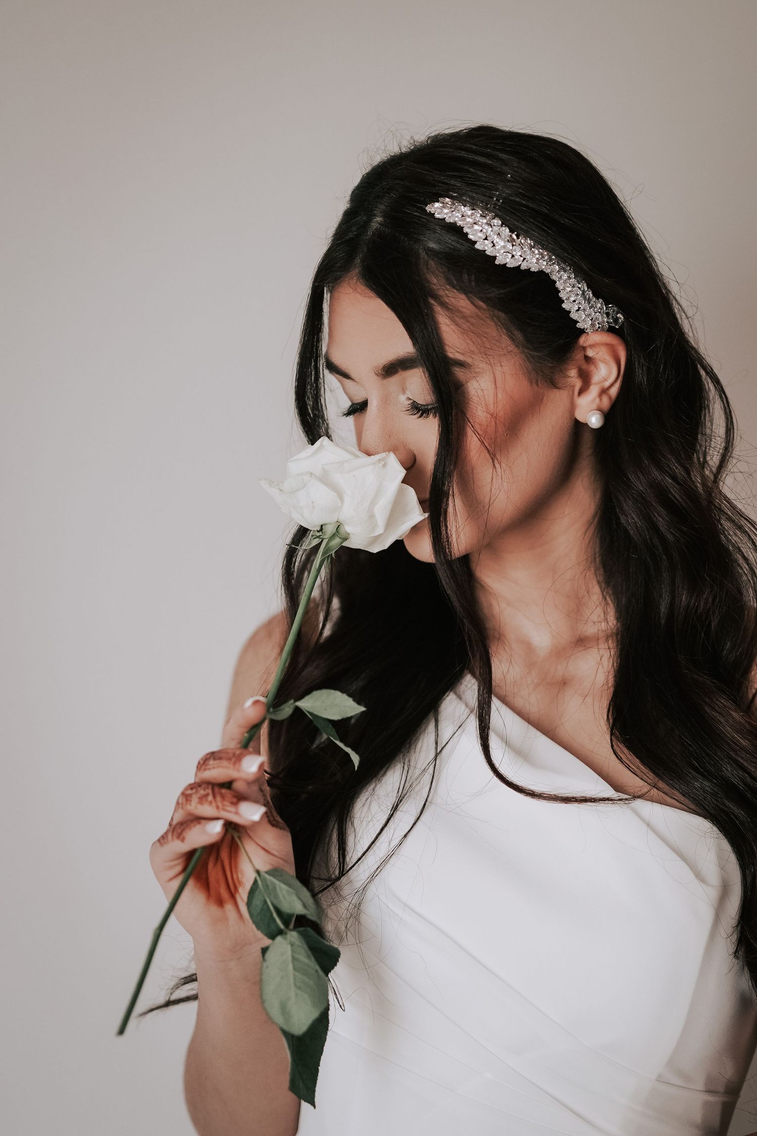 Dark-haired bride in white dress smells a single white rose while wearing a crystal hair accessory.