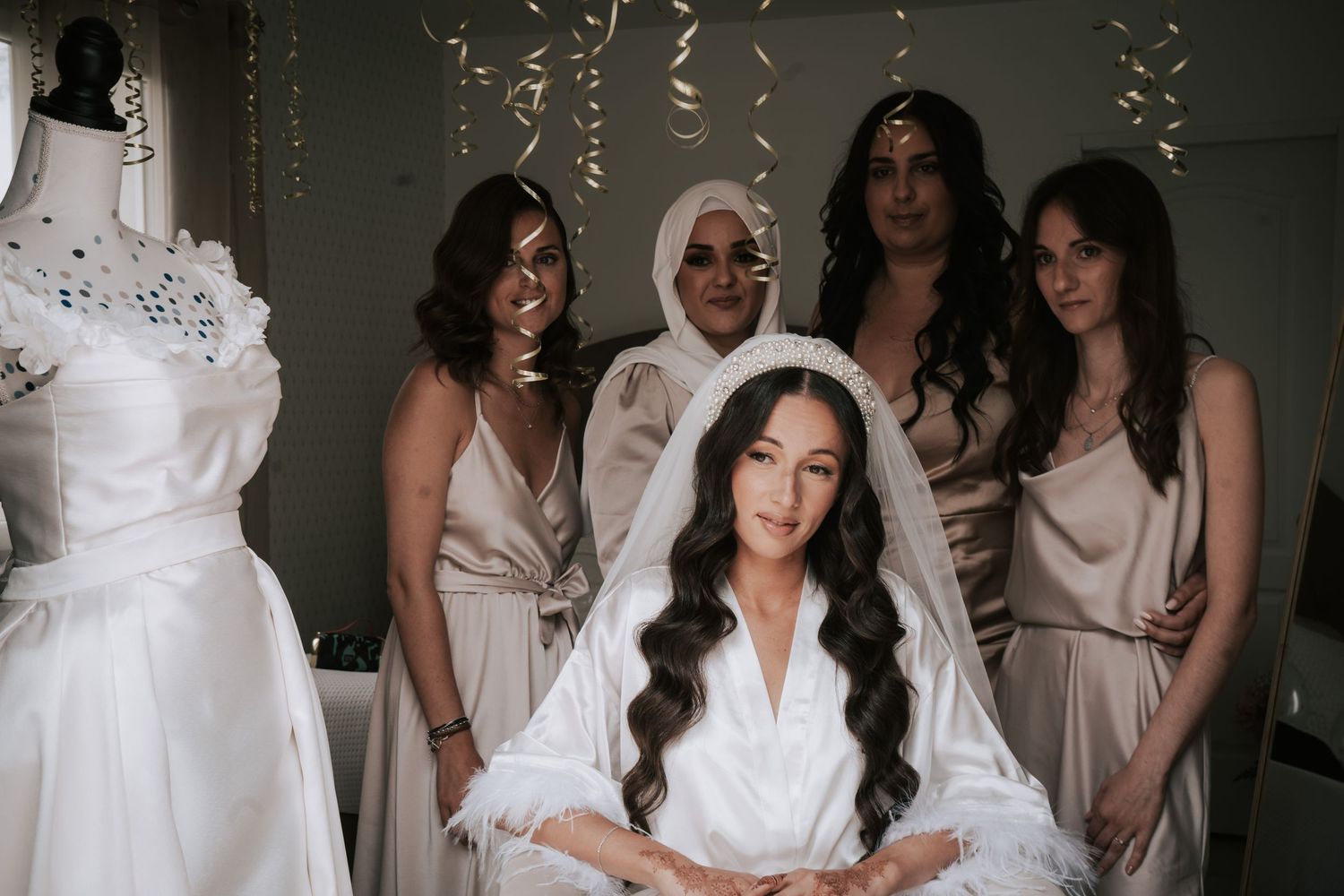Bride in white robe and veil poses with bridesmaids in champagne dresses in bridal suite.