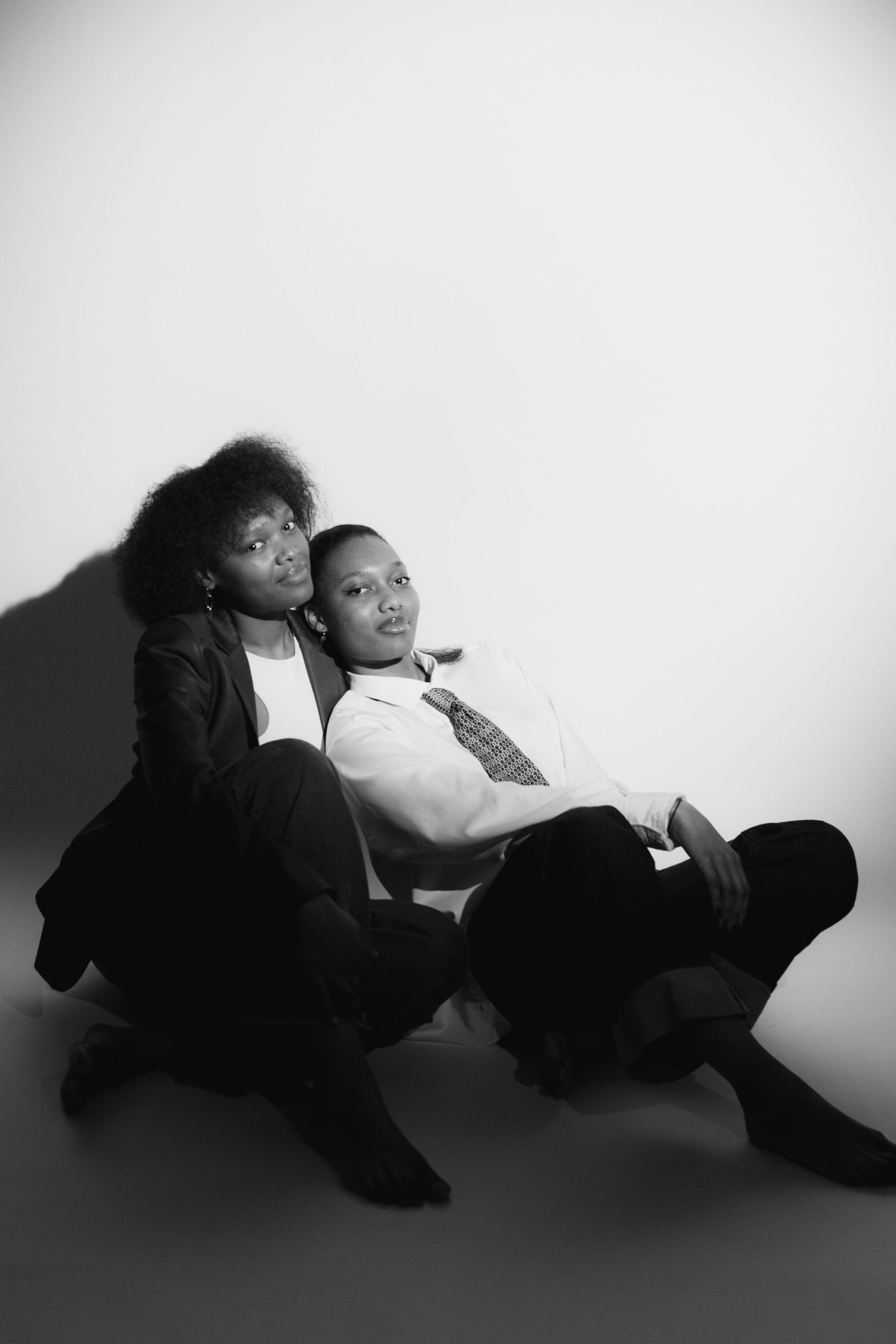 Black and white portrait of two people sitting together against a white wall, wearing formal attire.