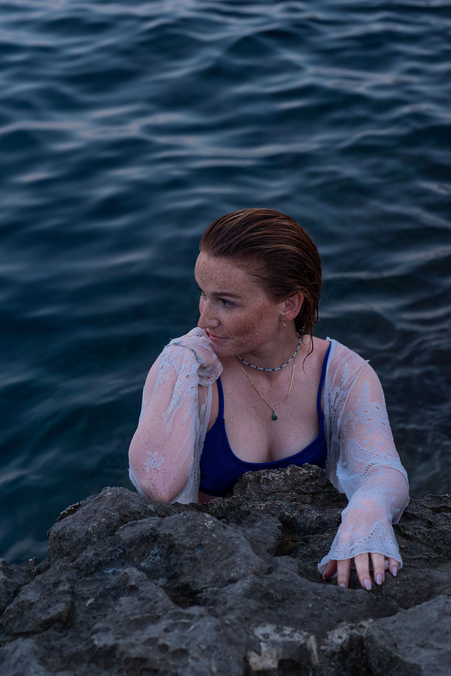 Freckled woman in blue swimsuit and sheer white cover-up posing on coastal rocks by the sea.