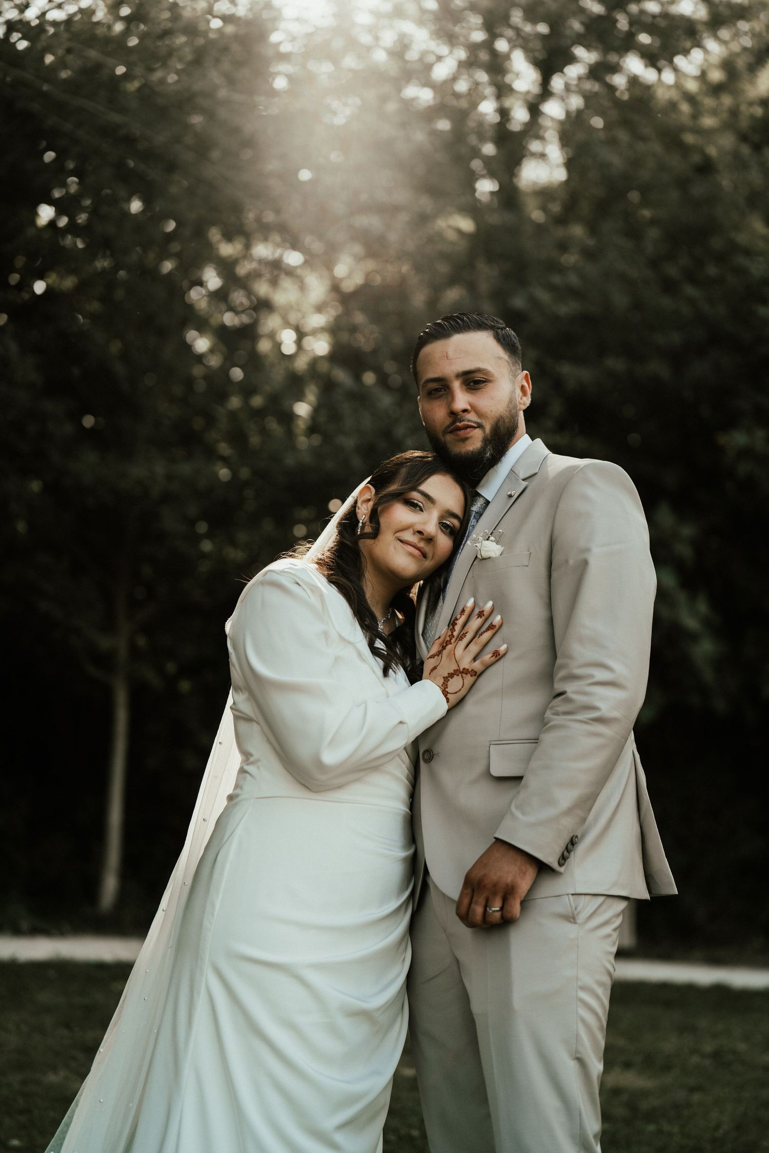 Bride leaning on groom showing wedding rings, embracing in a lush green garden at golden hour.