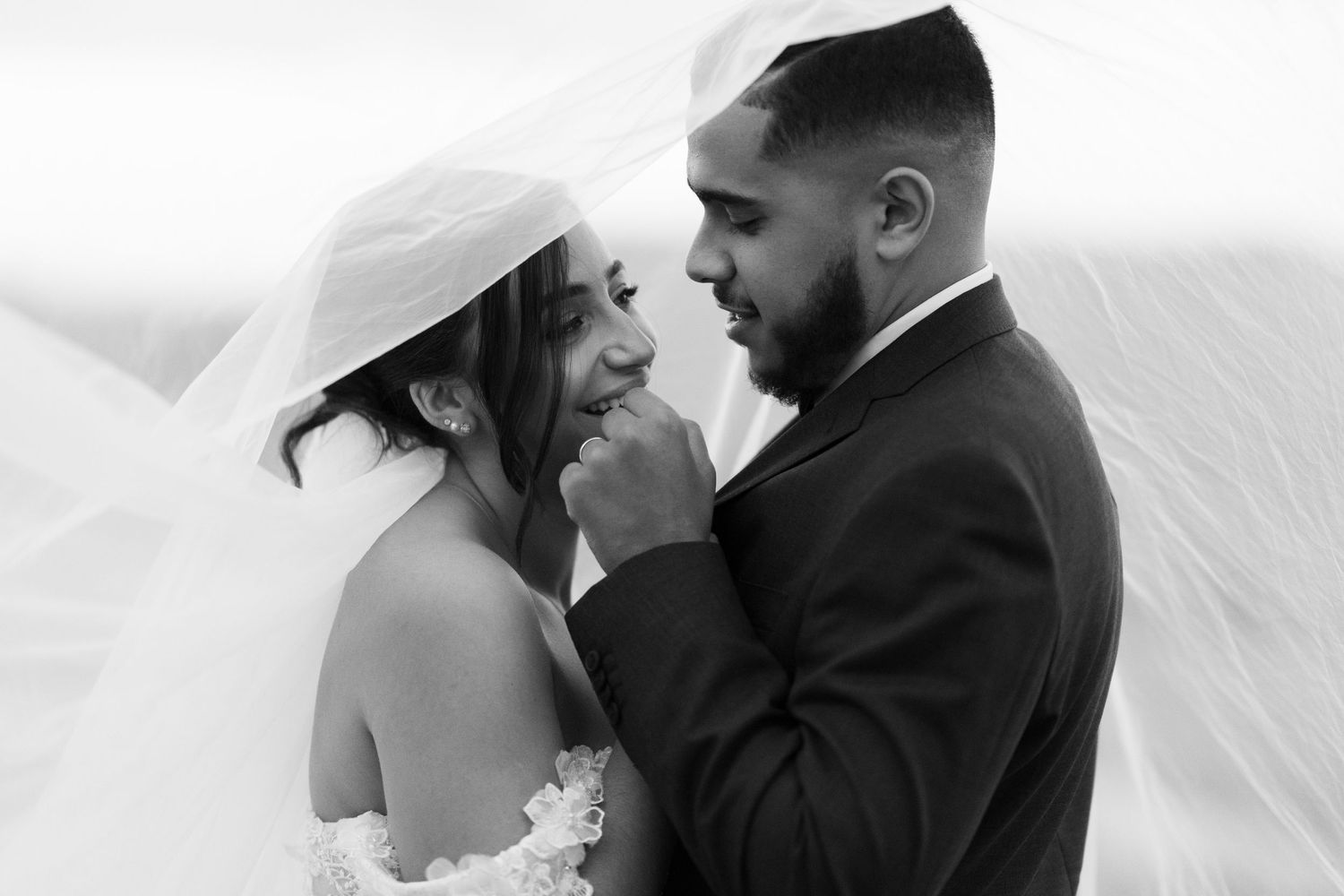 Black and white photo of groom tenderly holding brides face under flowing veil in romantic portrait.
