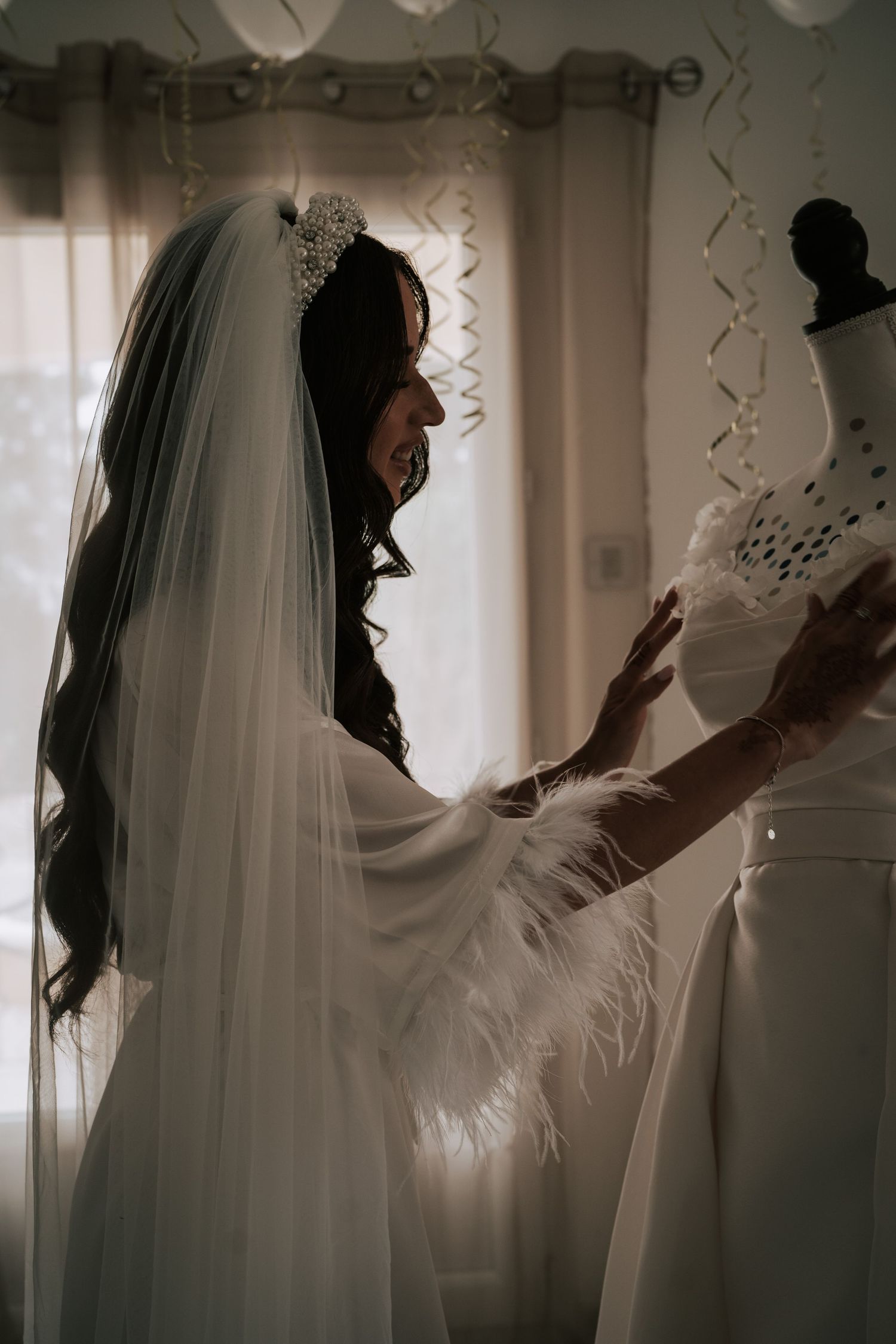 Bride in white veil and crown adjusts her wedding dress on a mannequin by a window.