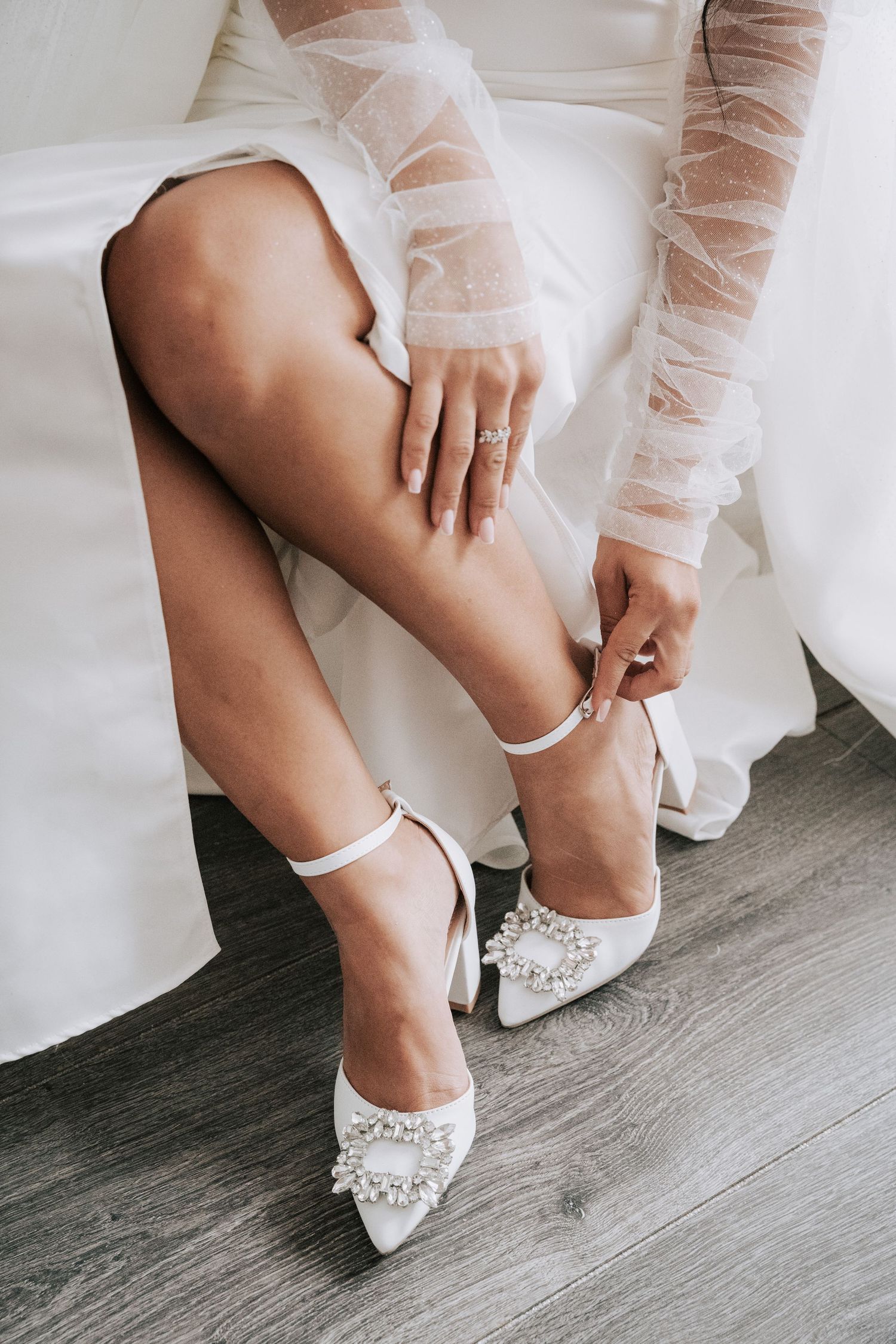 Bride fastening white pearl-embellished heels with ankle strap while wearing lace wedding dress.