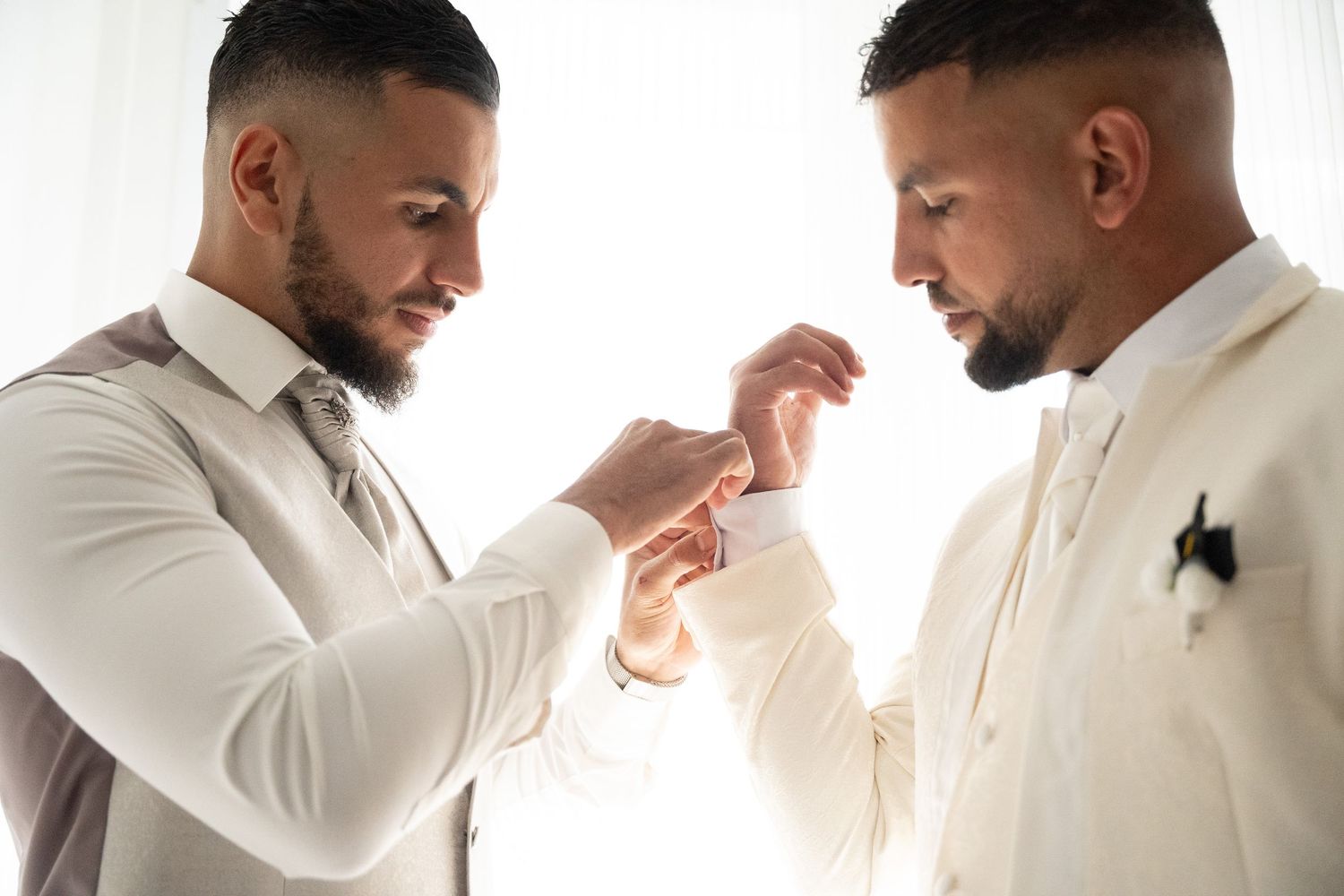 Groom in white suit having cufflinks fastened by bearded groomsman before wedding ceremony.