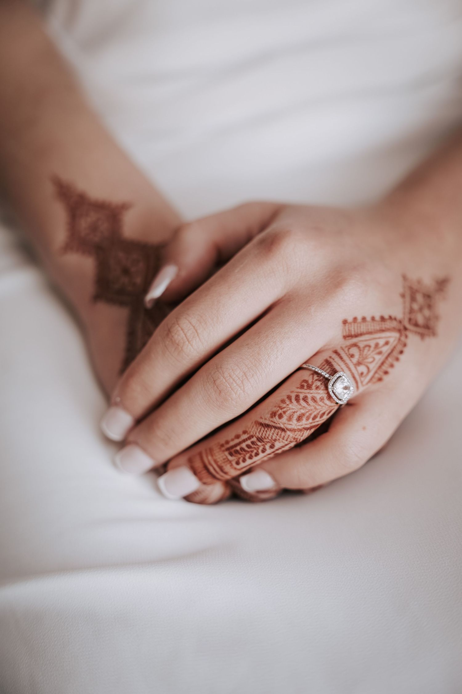 Bride's hands with henna tattoos and a diamond engagement ring resting on white wedding dress.
