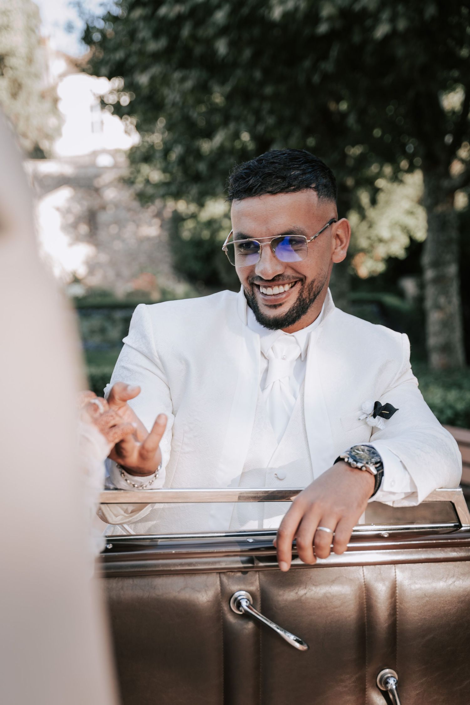 Smiling groom in white suit with glasses gestures beside a vintage car in sunny garden.
