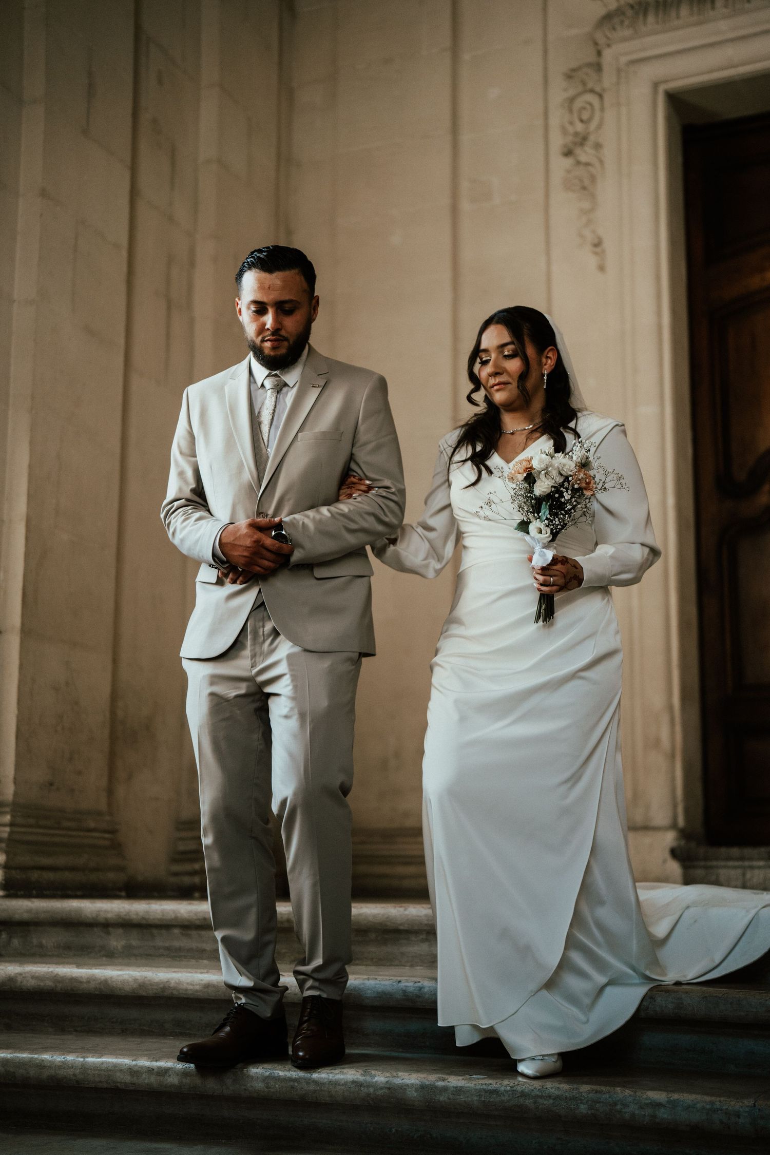 Bride and groom standing on stone steps outside a grand building on their wedding day.