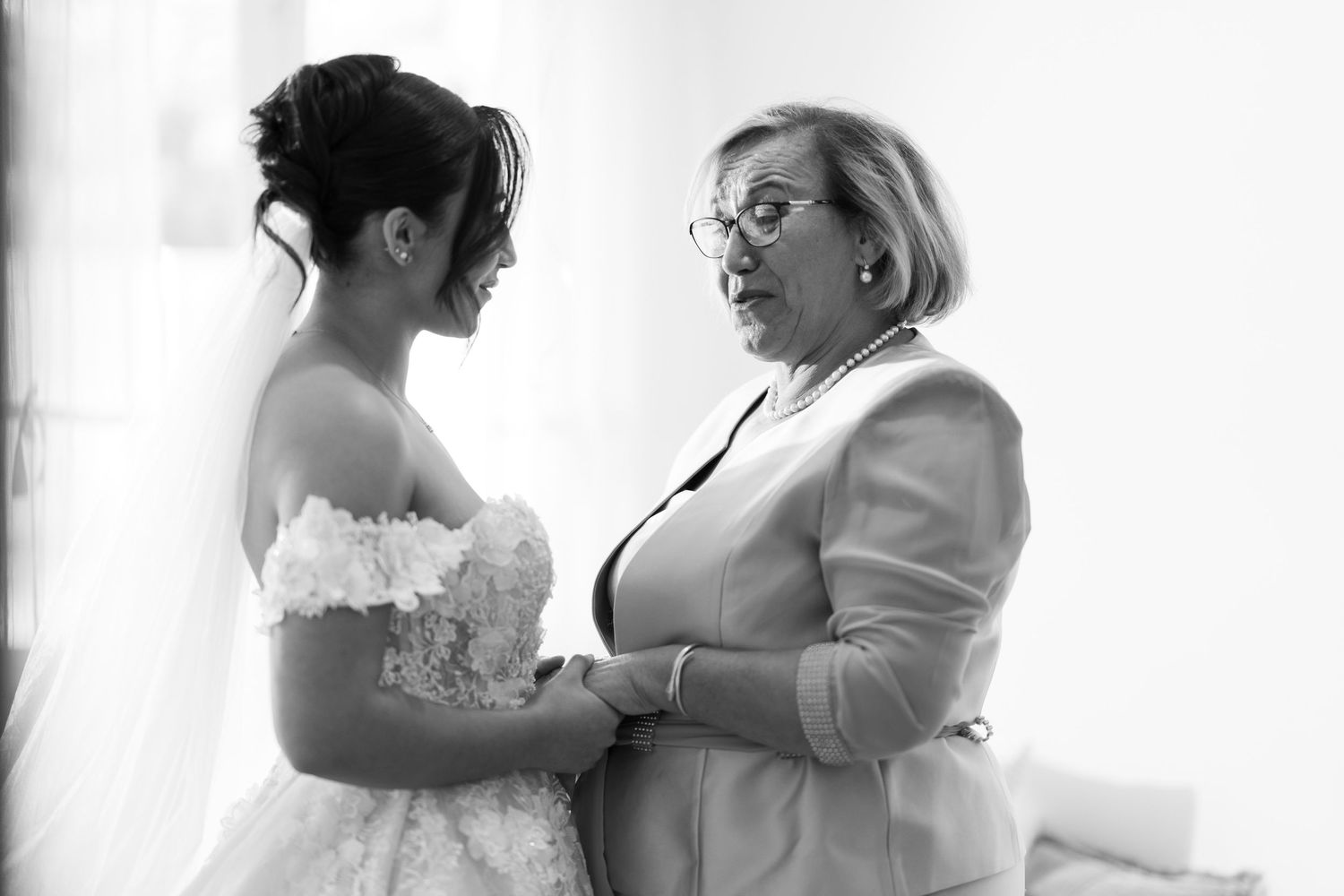 Bride in lace wedding gown holding hands with older woman in black and white photo.