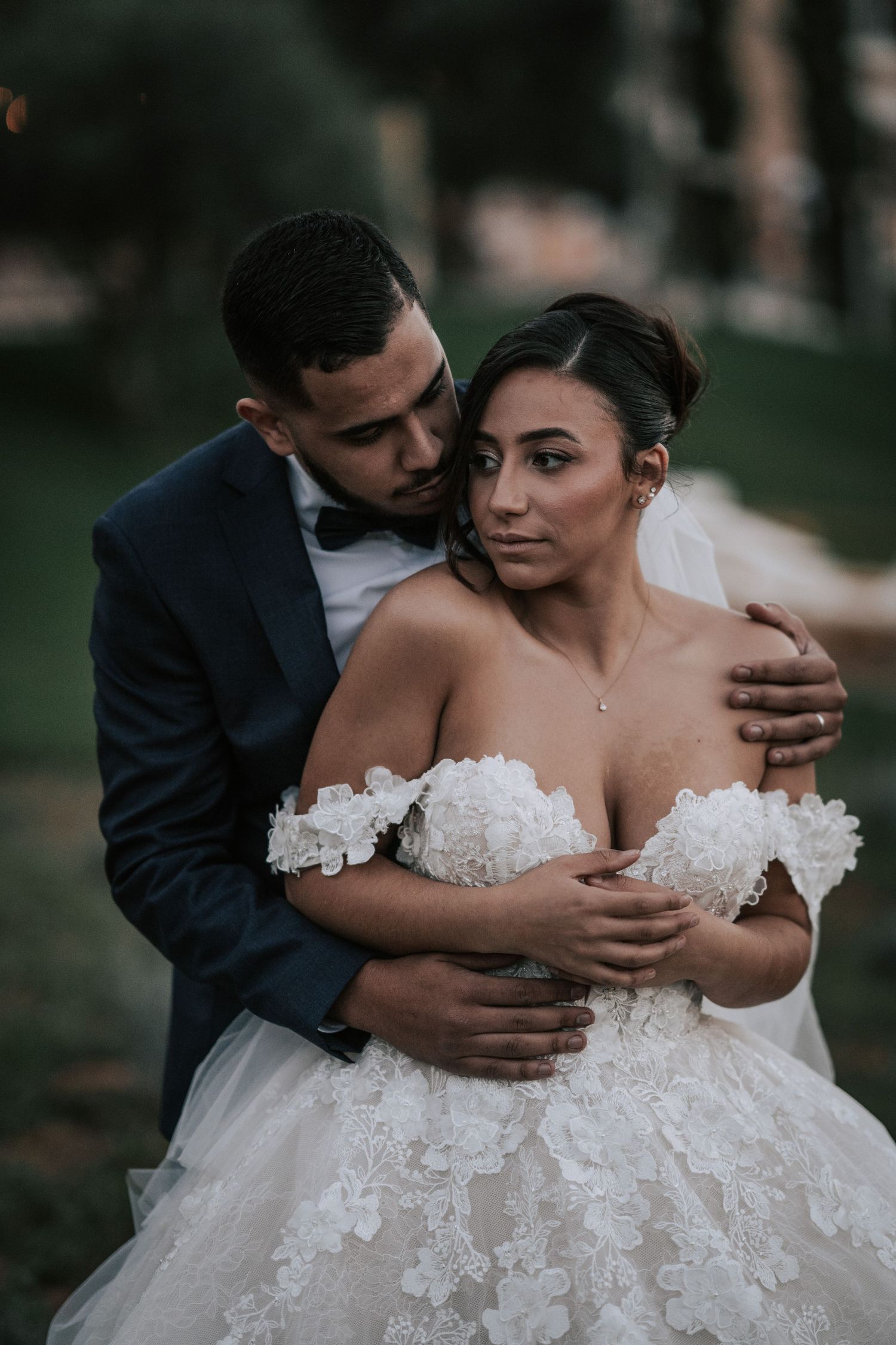 Groom in navy tuxedo embraces bride from behind in floral lace ballgown in a romantic outdoor portrait.