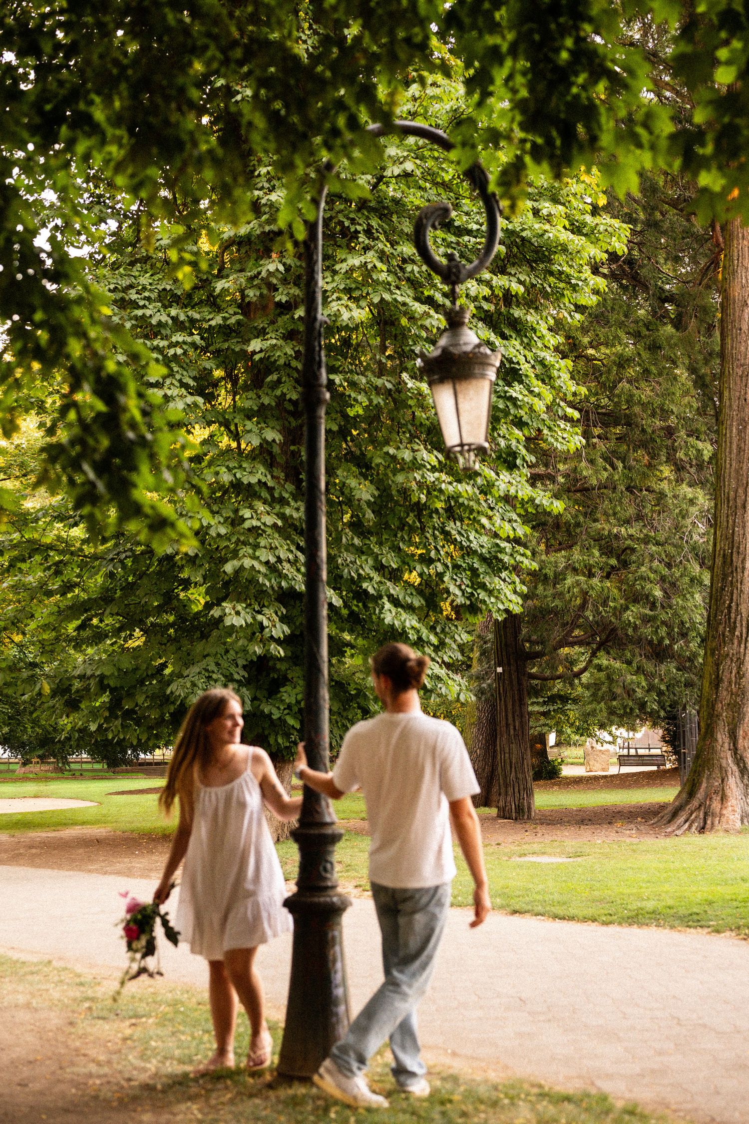 Couple walking through a lush green park, woman in white dress holding flowers, passing an ornate street lamp.