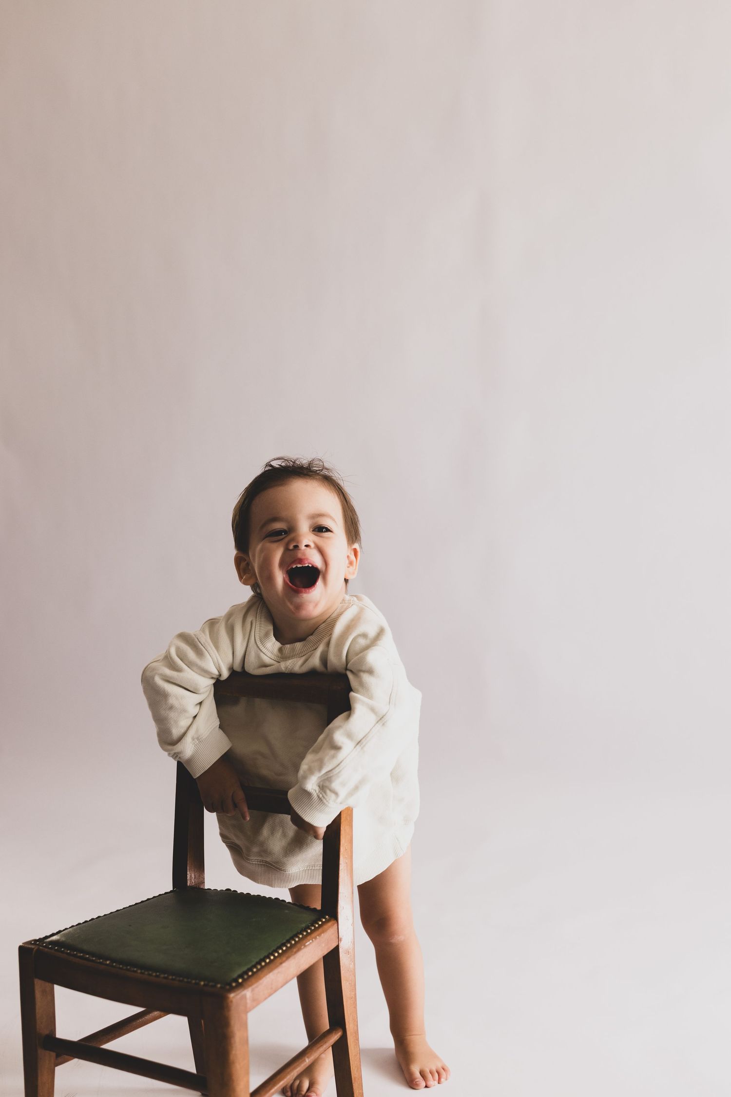 An excited laughing toddler in a cream romper leans over a vintage wooden chair in a bright studio.