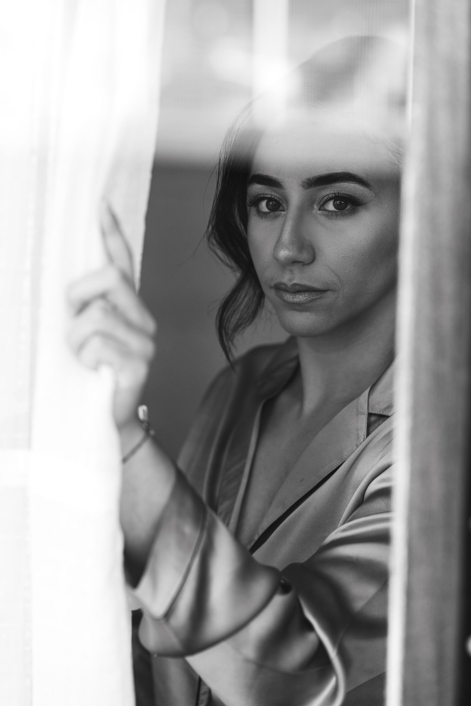 Black and white portrait of bride in satin robe holding sheer curtain by window.