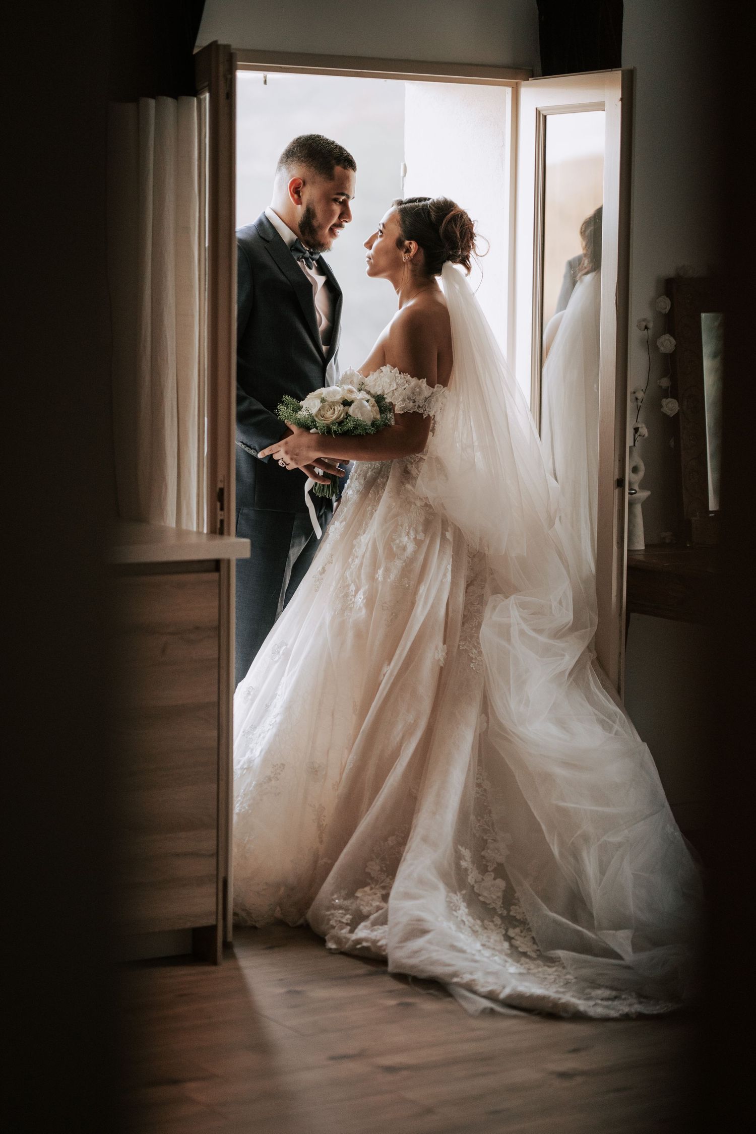 Bride and groom share a tender gaze in elegant doorway, bride holding white floral bouquet.