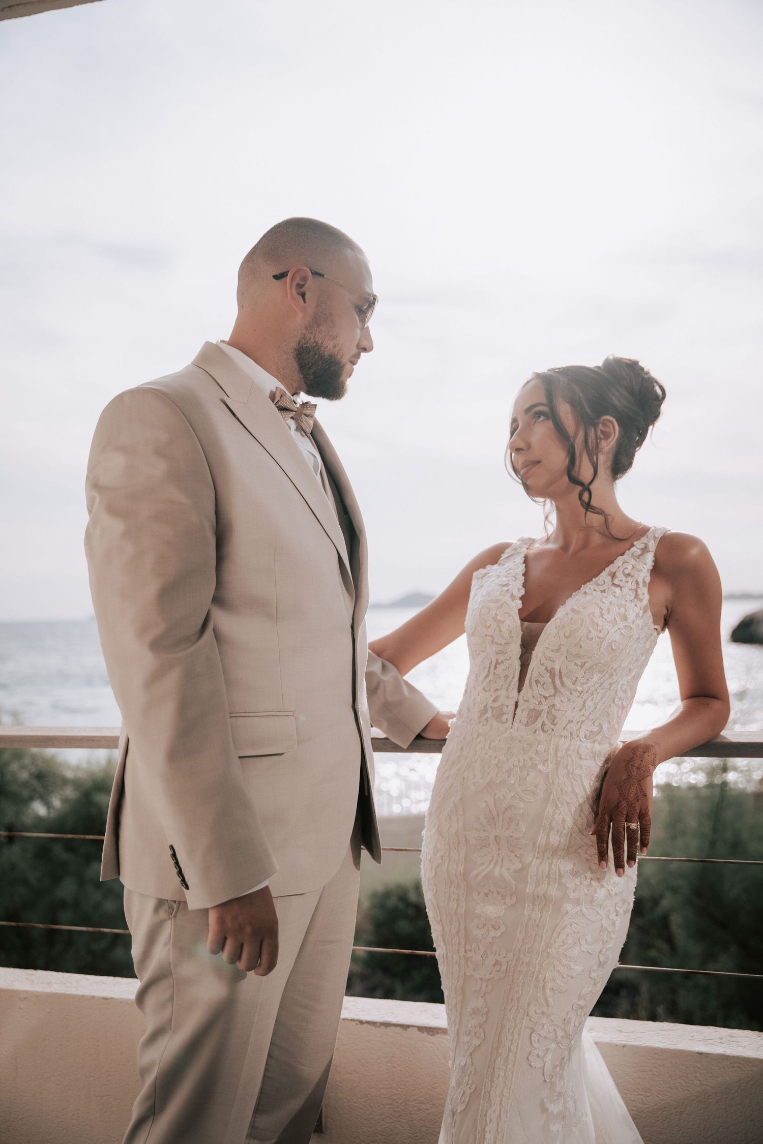 Bride and groom in beige suit share romantic moment on oceanview balcony.