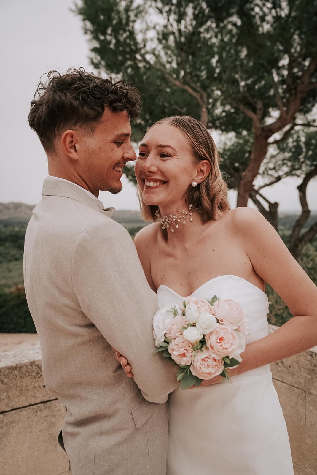 Joyful bride in strapless white gown laughs with groom in beige suit holding pink peony bouquet.