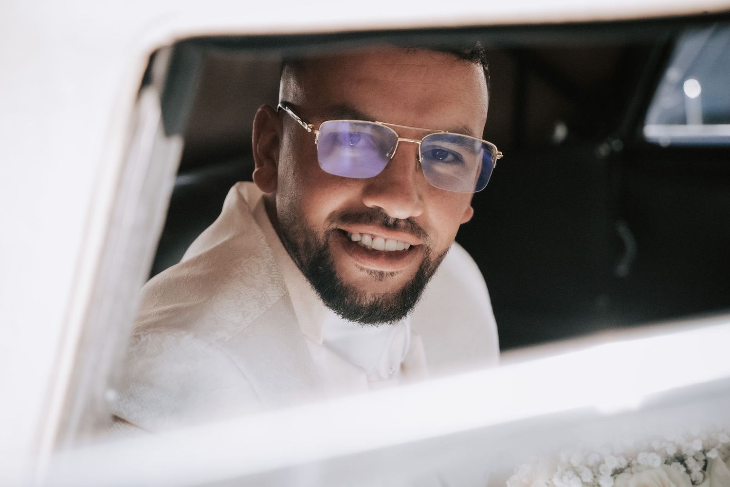 Smiling groom with glasses peers through a car window on his wedding day.