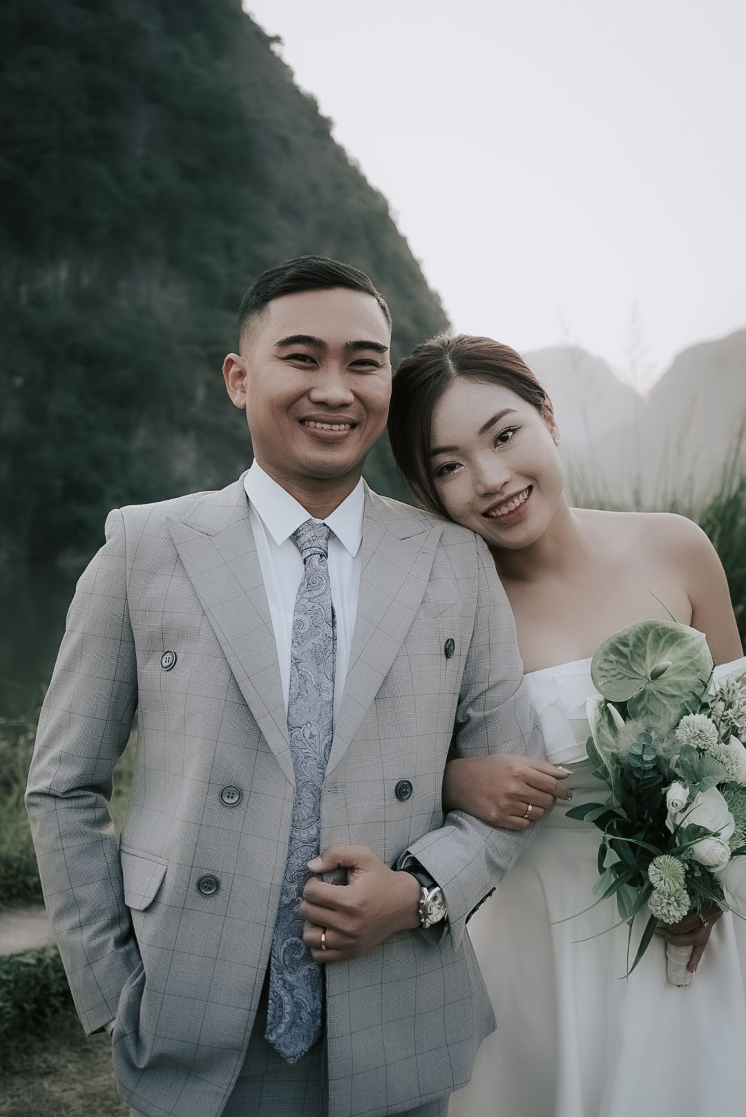 Happy Asian bride and groom in grey suit pose together with greenery bouquet outdoors.