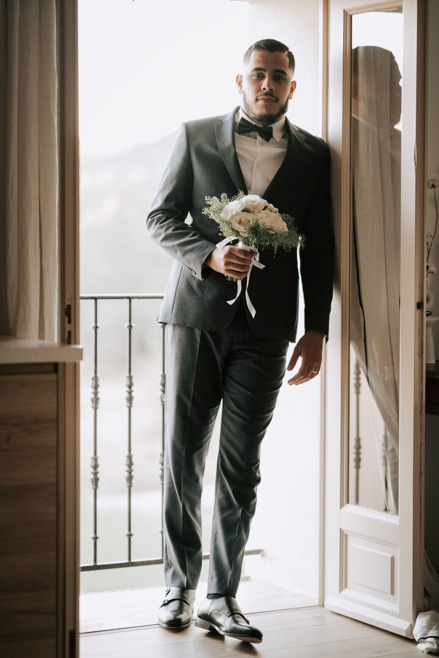 Elegant groom in gray suit holds white bouquet standing in doorway with balcony.