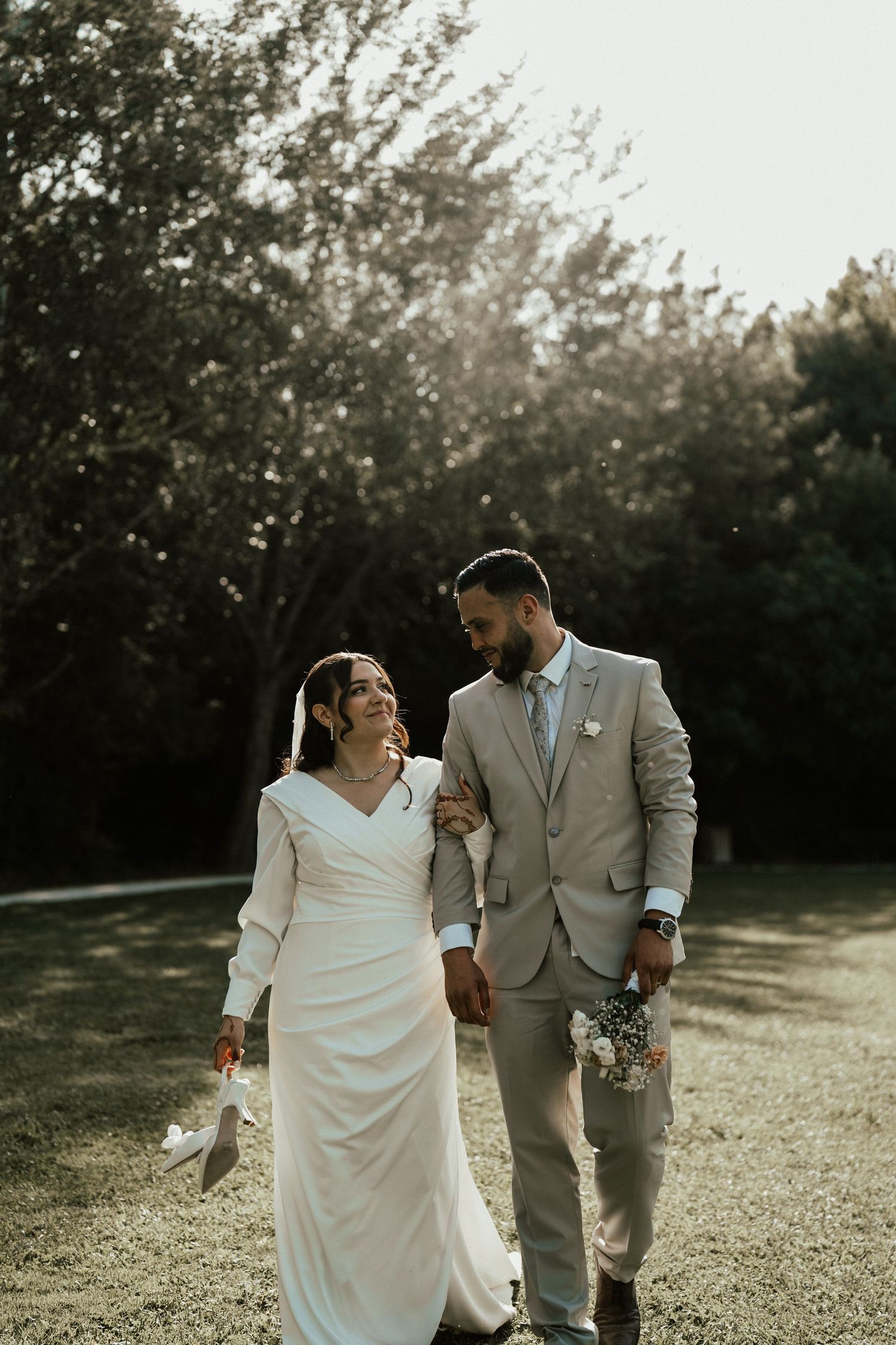 Bride and groom walking hand in hand through a sunlit garden, smiling at each other on wedding day.