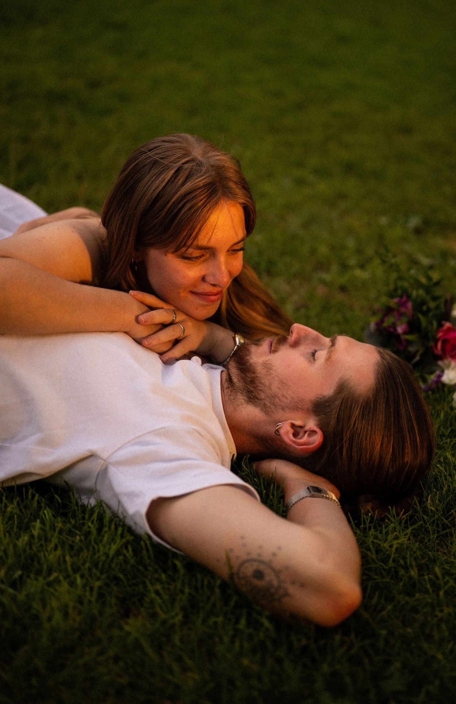Romantic couple lying on green grass, woman smiling while resting on mans chest outdoors.