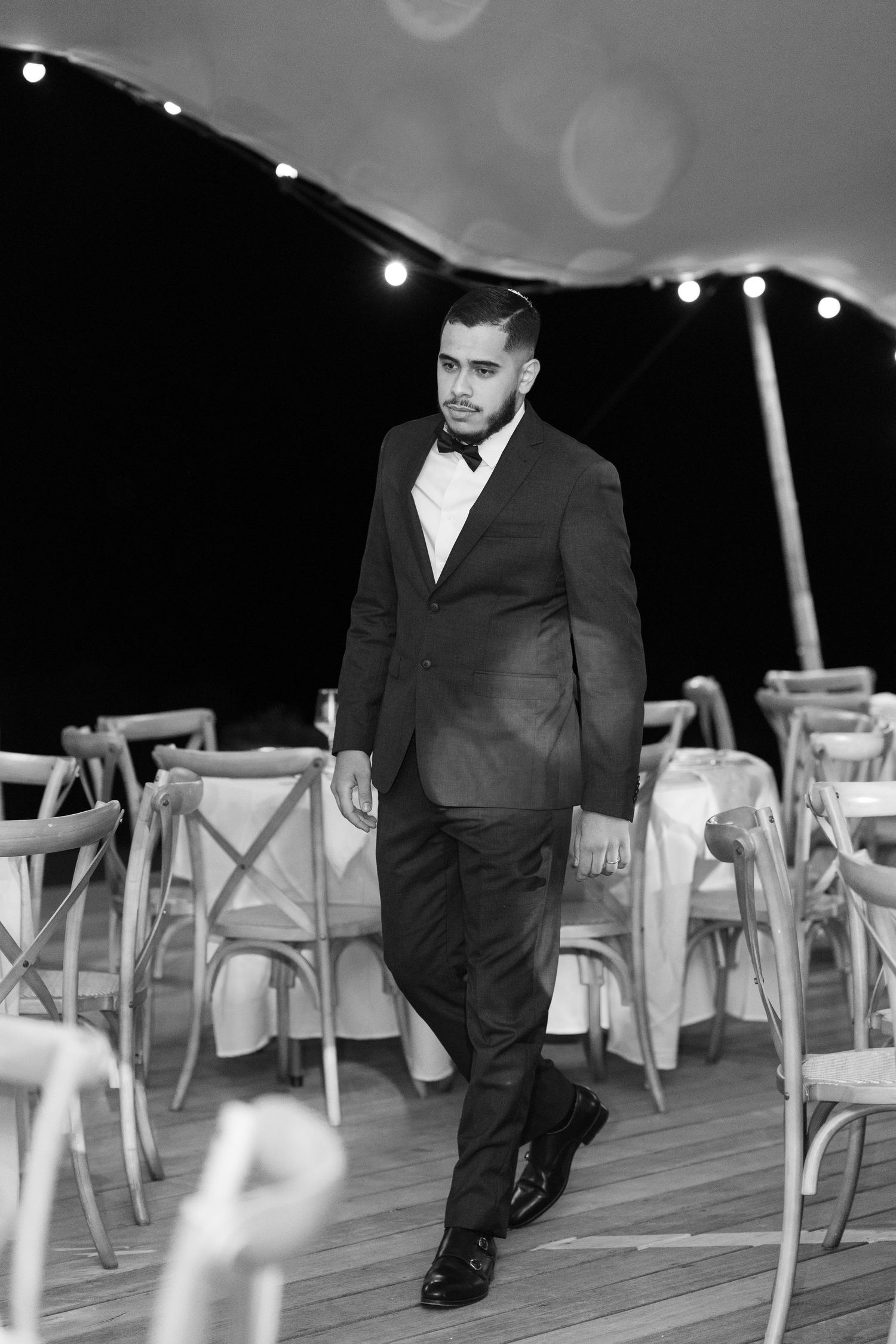 Dapper groom in a black tuxedo and bow tie walking confidently through an elegant tented wedding venue.