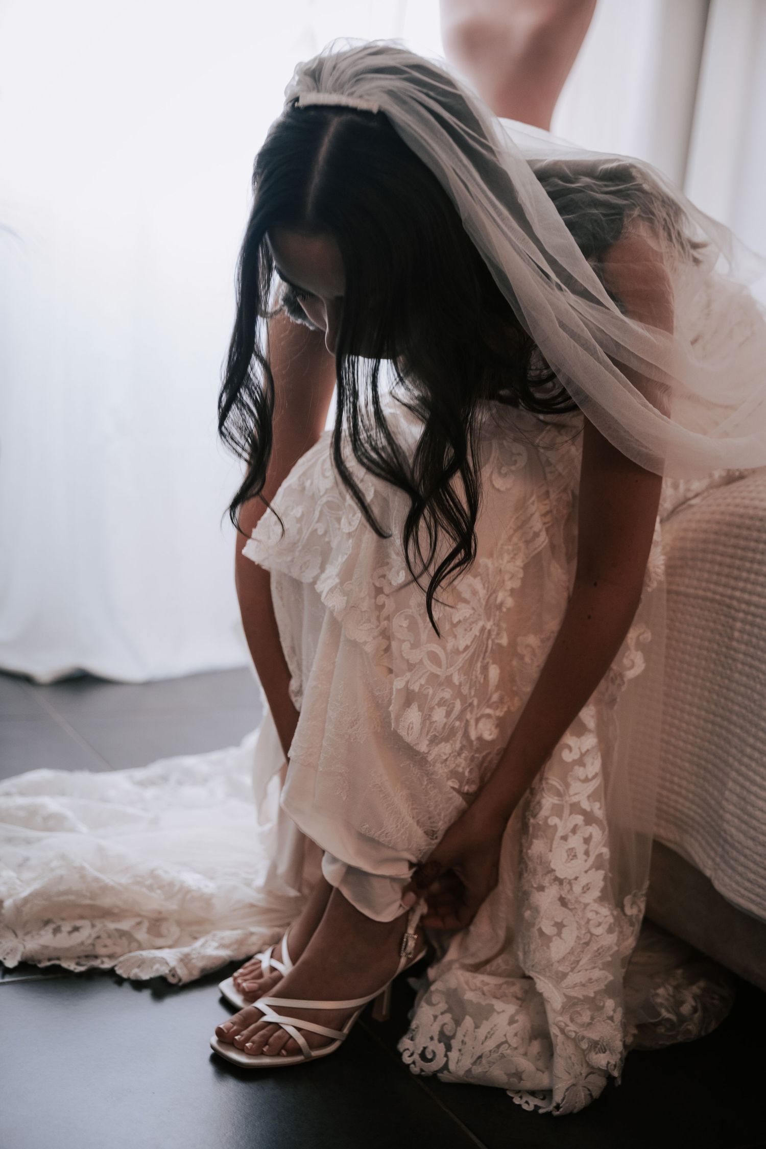 Bride in lace wedding gown and veil putting on white strappy heels before the ceremony.