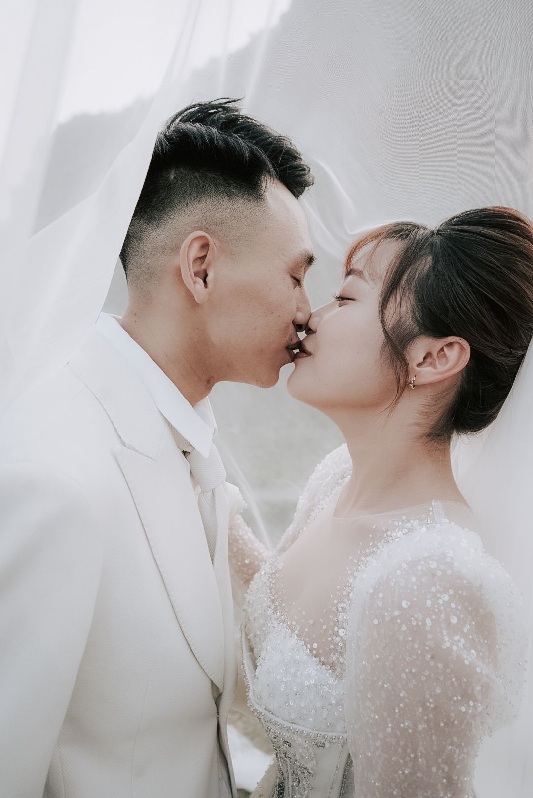 A couple in white wedding attire shares a romantic kiss under a flowing veil in an elegant bridal portrait.