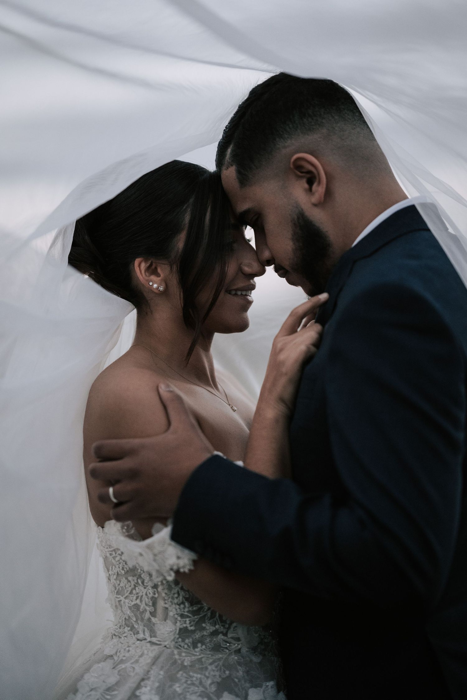 Bride and groom share an intimate moment under a billowing veil, smiling and touching foreheads tenderly.
