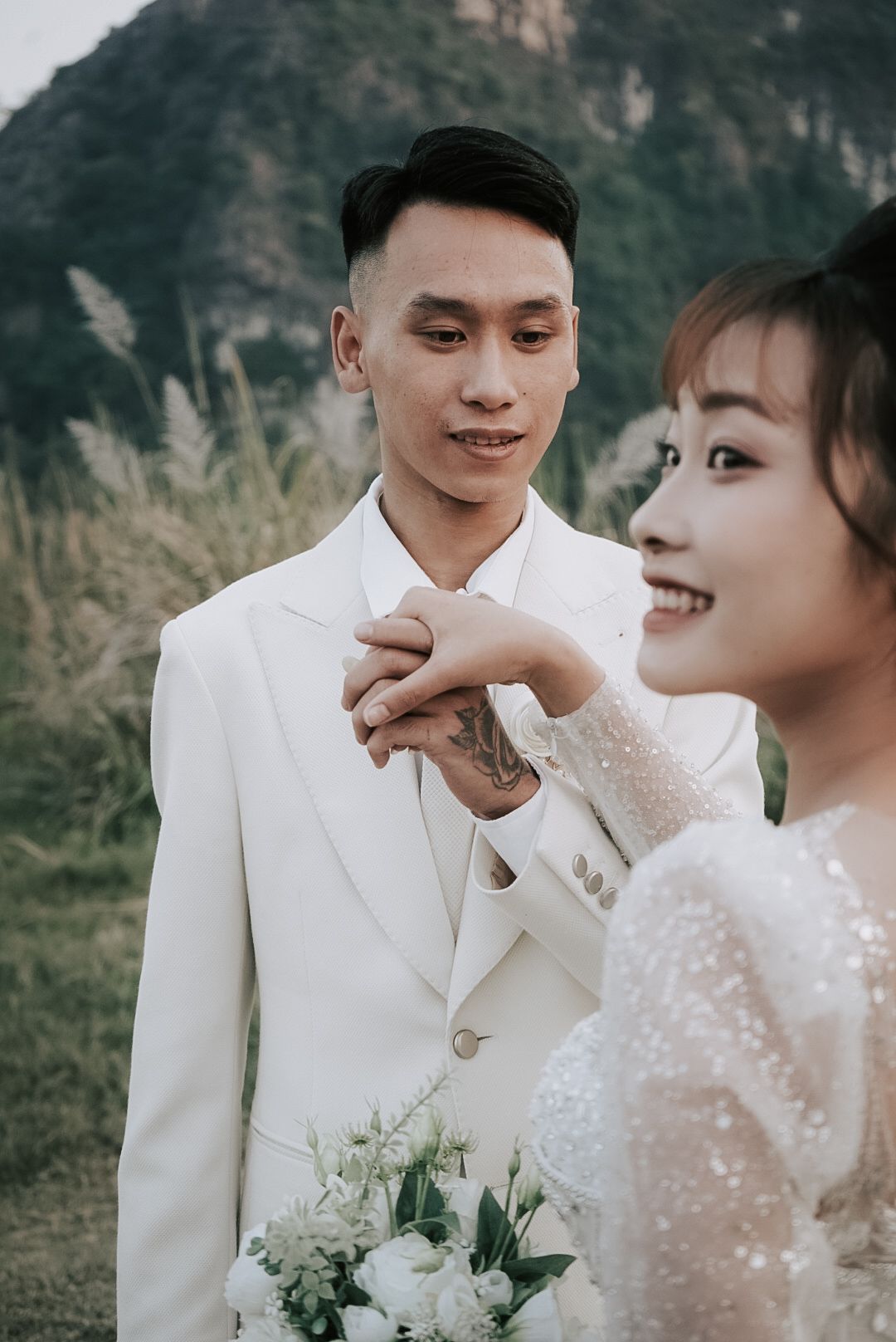 Groom in white suit tenderly holds bride's hand outdoors with mountains in background.