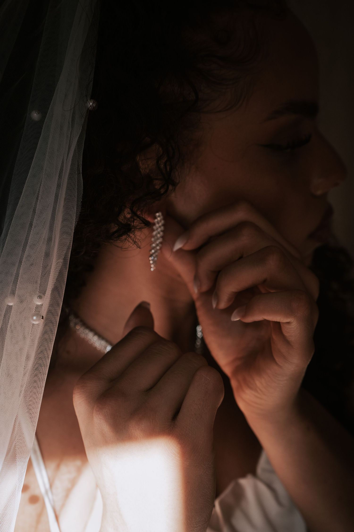 Close-up of a bride with curly hair putting on a dangling crystal earring beneath a pearl-trimmed veil.