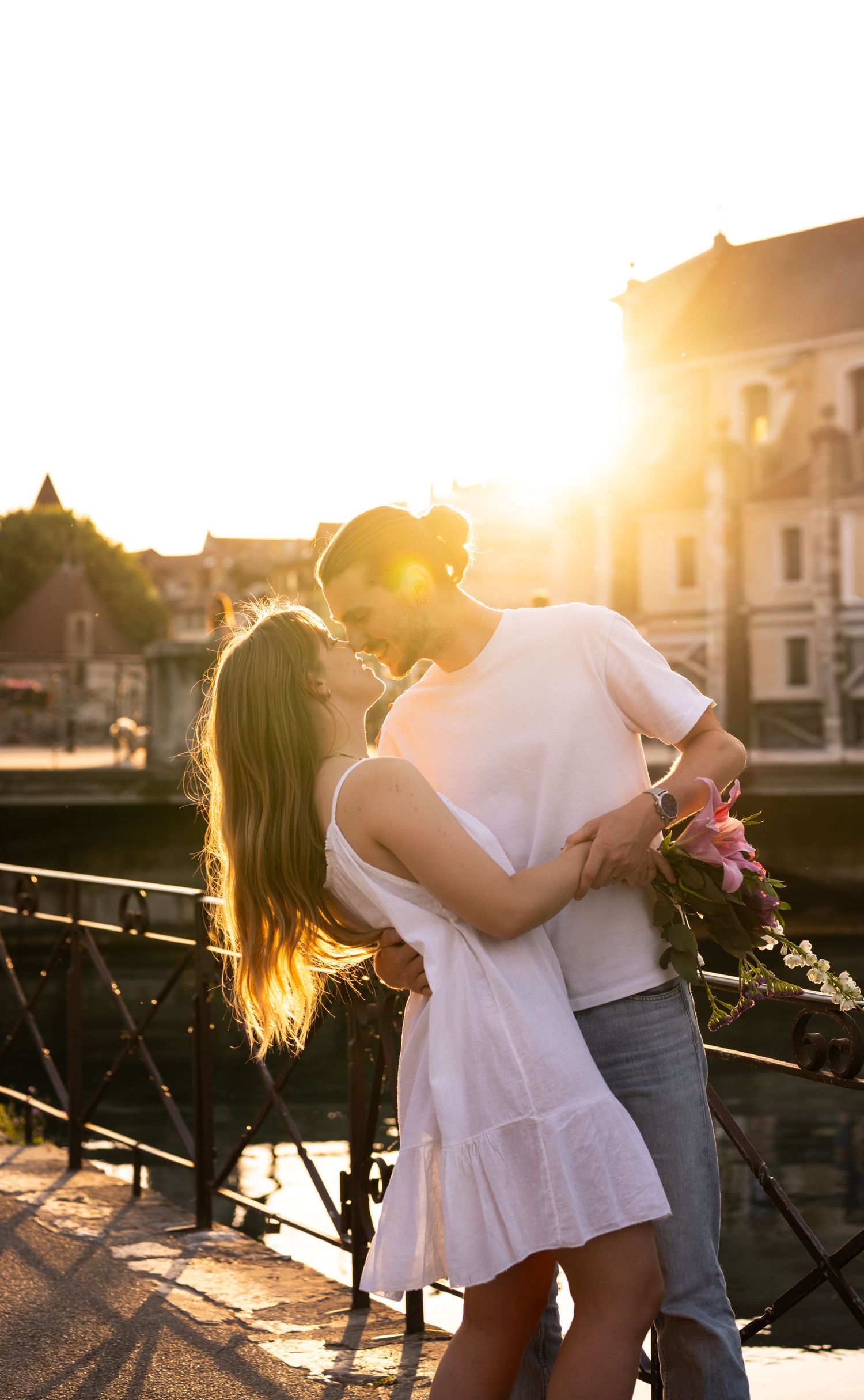 Couple kissing on a bridge at golden sunset, woman in white dress holding pink flowers.