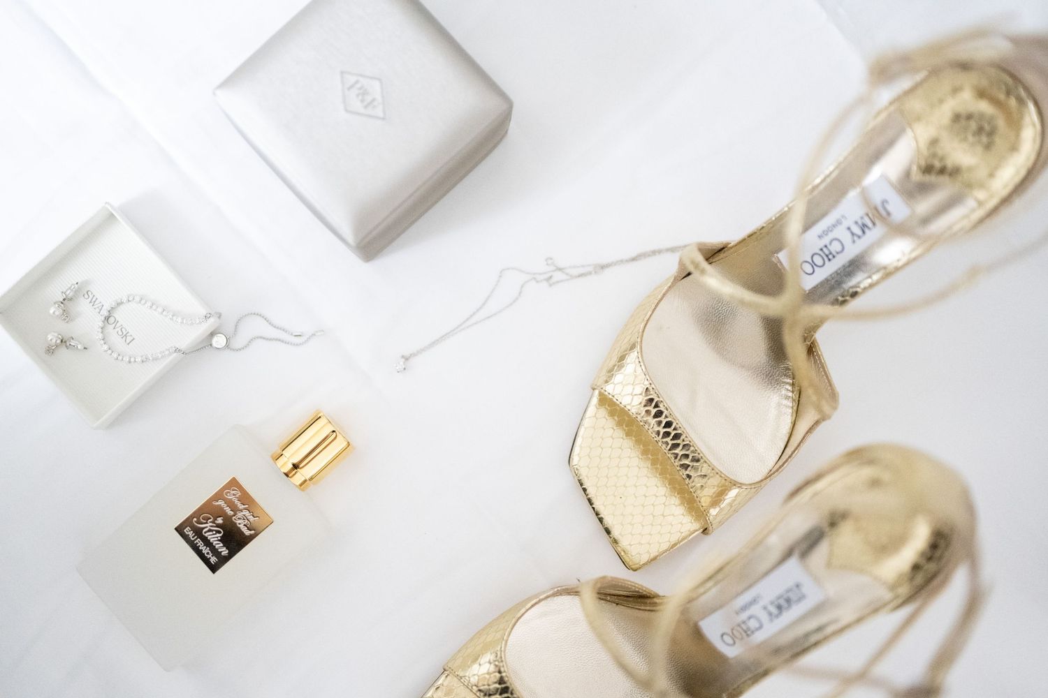 Gold Jimmy Choo heels, perfume, diamond necklace, and jewelry box arranged elegantly on white surface.