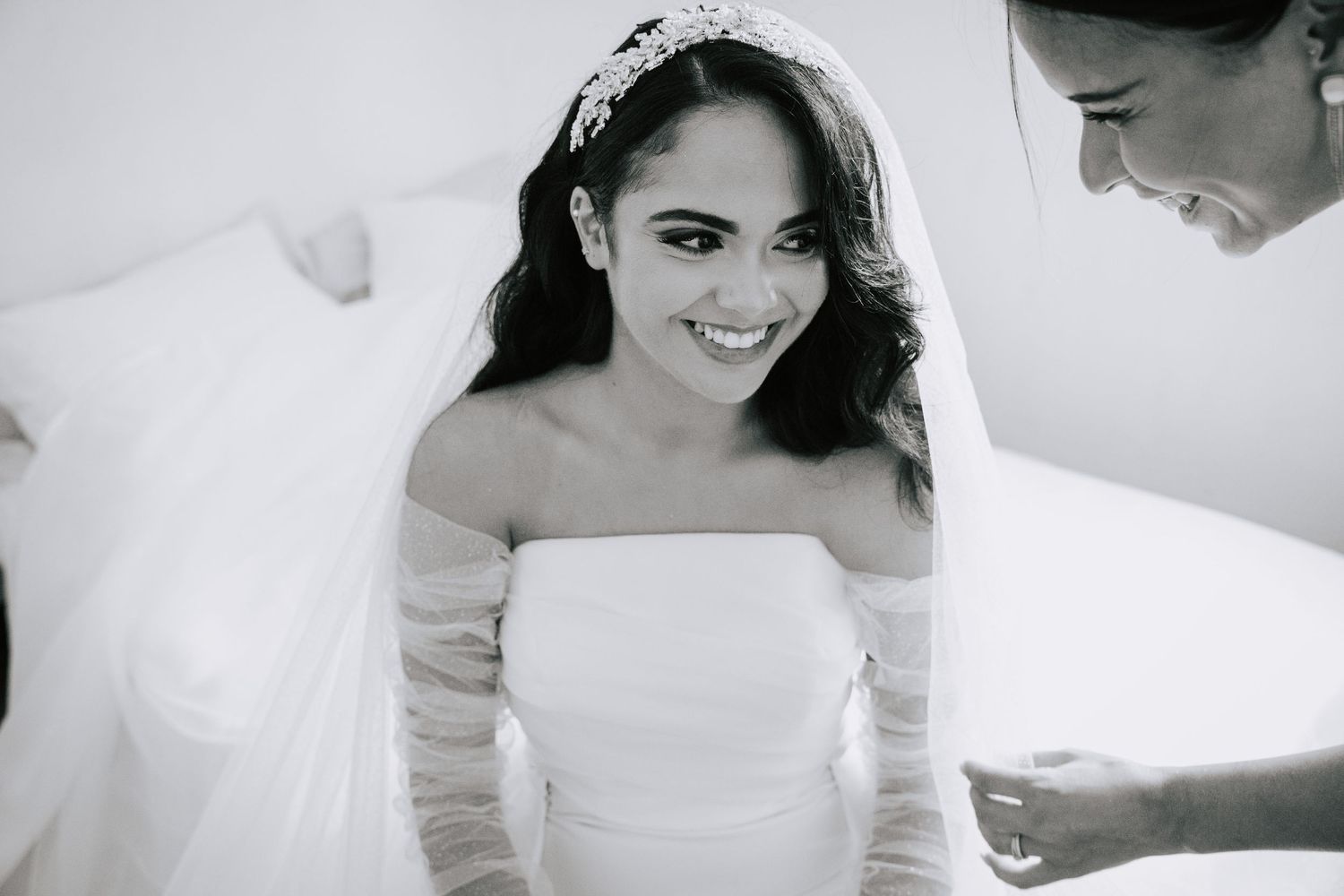 Smiling bride in white off-shoulder gown and floral headpiece is assisted with her dress in black and white.