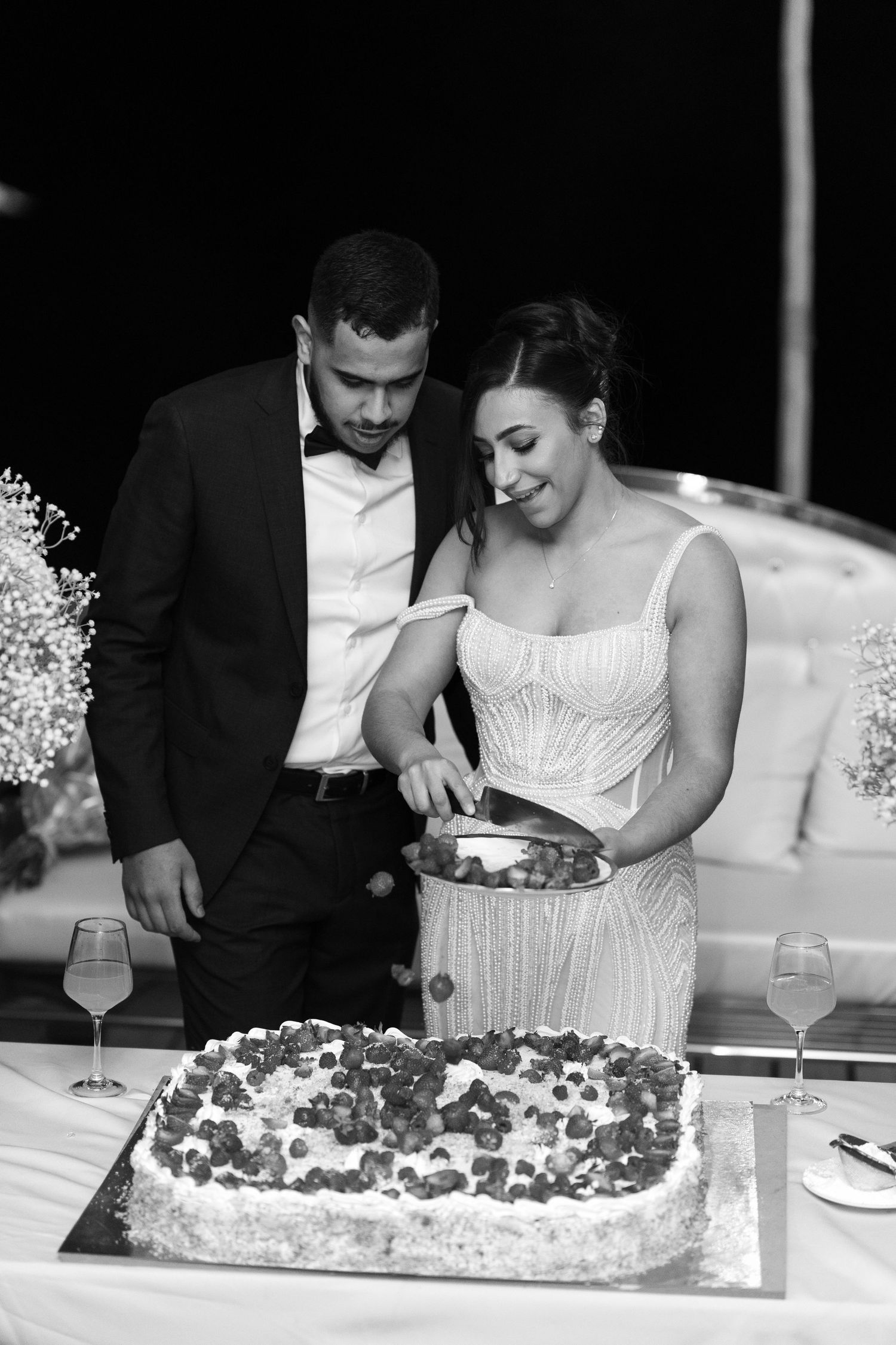 Black and white photo of bride and groom cutting large berry-topped wedding cake at outdoor reception.