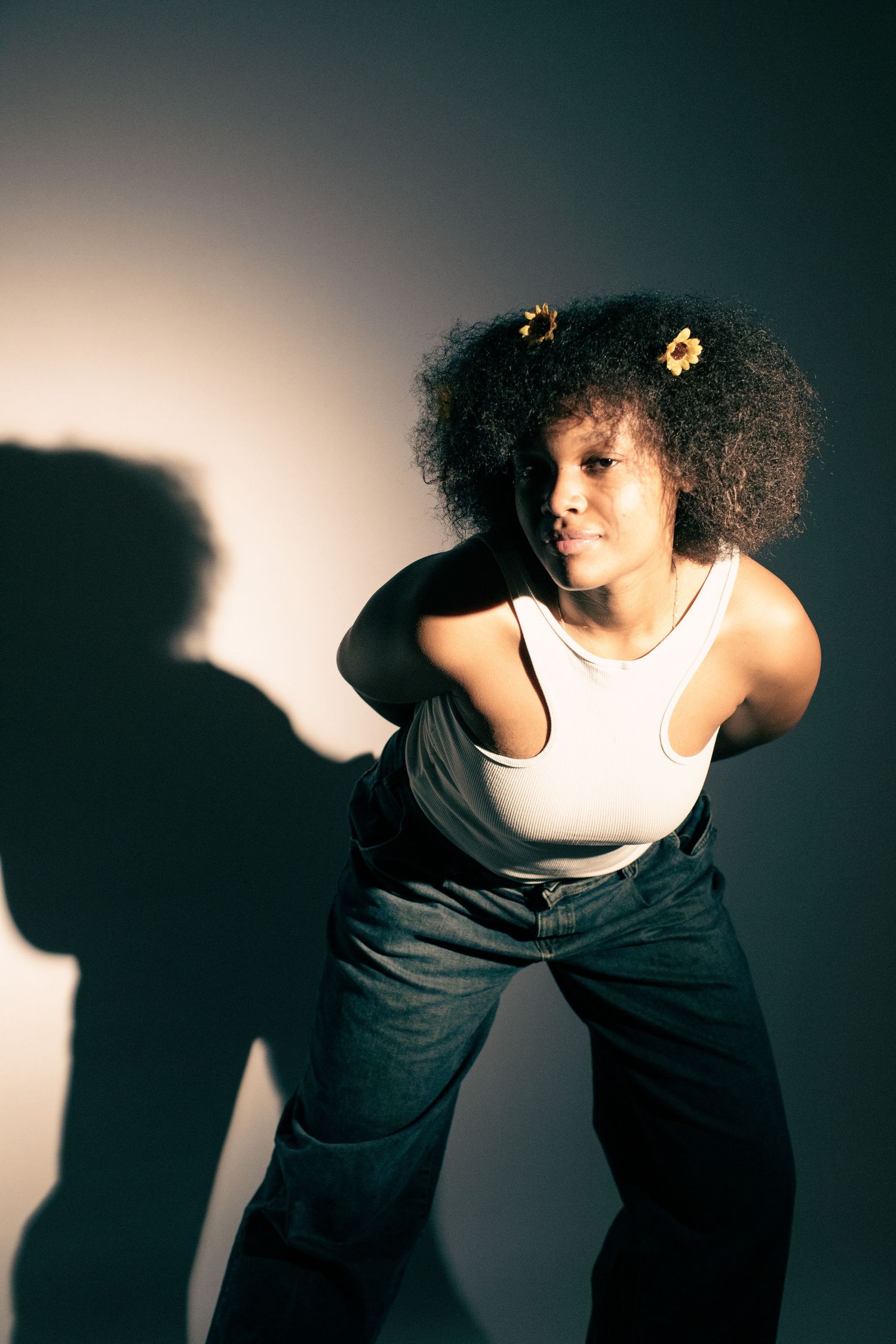 Young Black woman with afro and sunflower hair clips poses confidently in white tank top and jeans against gray background.