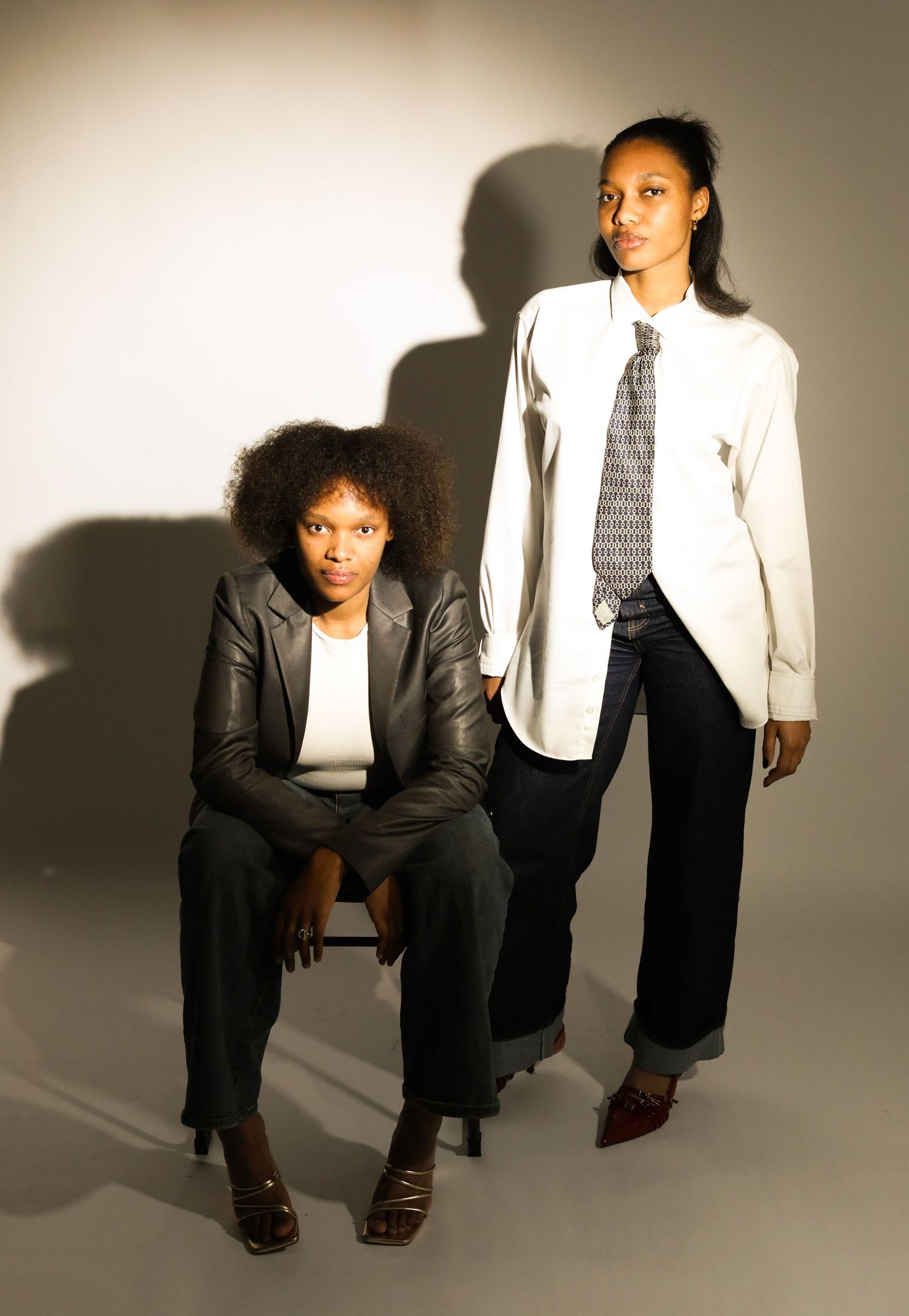 Two stylish Black women posing for a portrait, one seated in a blazer, one standing in a white shirt and tie.