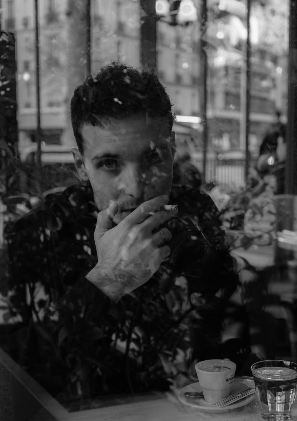 Black and white photo of a young man smoking a cigarette at a cafe table with coffee cups, shot through a window.
