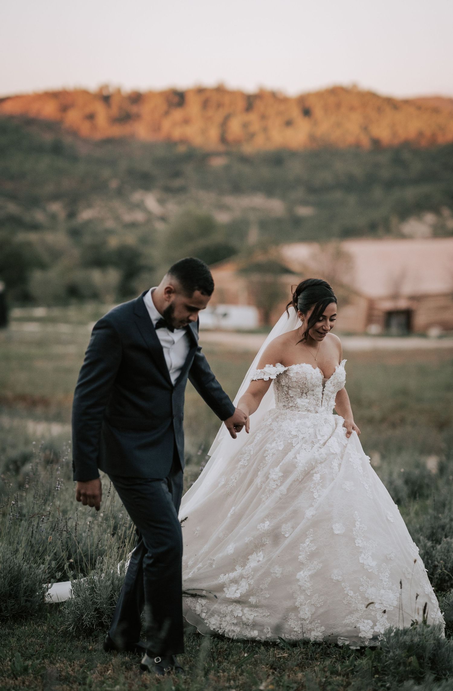Bride and groom walk hand in hand through golden field at sunset with scenic hills behind.
