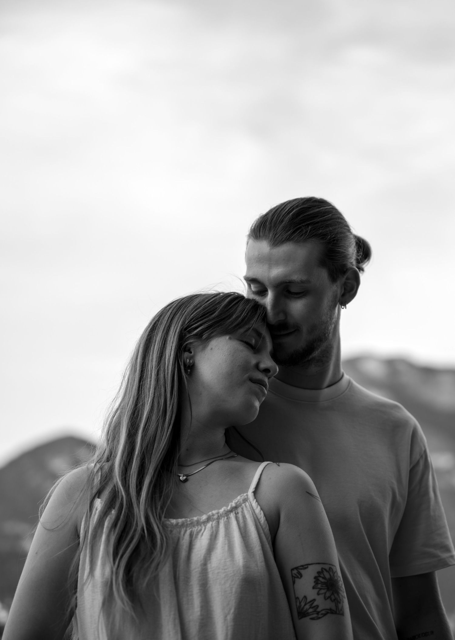 Black and white portrait of a couple outdoors, woman leaning into man with tattoo, mountains visible behind them.