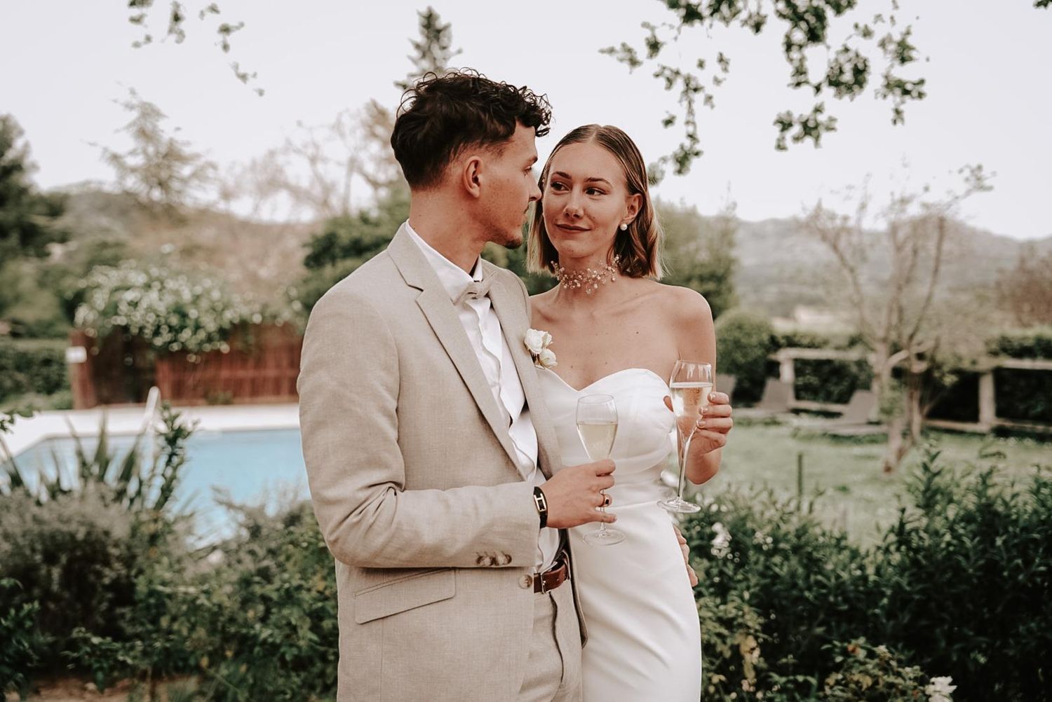 Newlyweds in elegant wedding attire toast with champagne glasses in a lush garden setting near a pool.