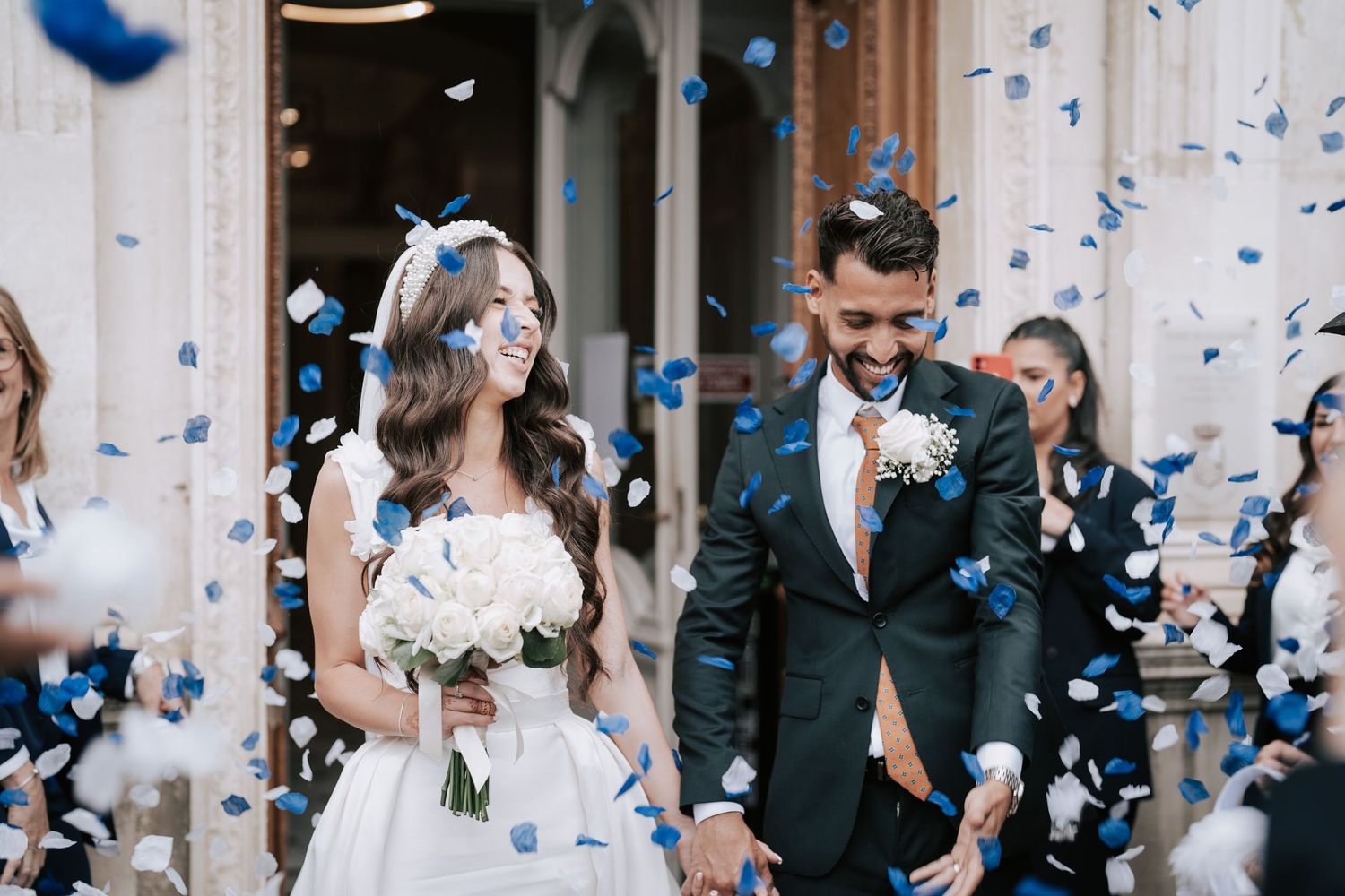 Happy newlyweds walk through blue and white confetti shower outside elegant venue on their wedding day.