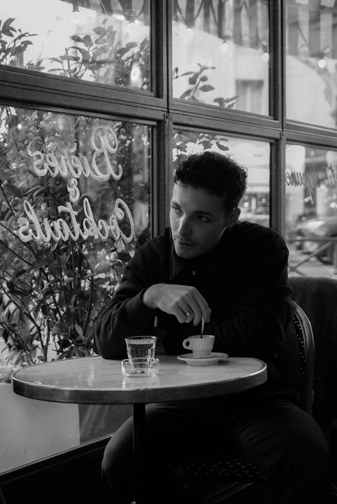 Black and white photo of a young man stirring coffee at a Parisian cafe table by a window.