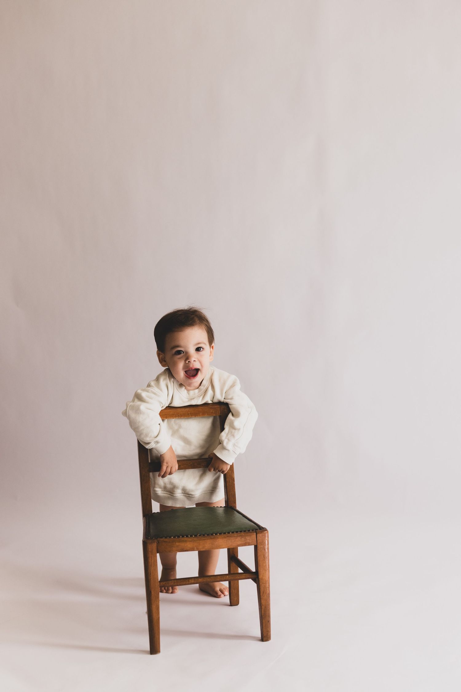 A joyful toddler in a cream romper stands at a vintage wooden chair with a green seat in a studio.