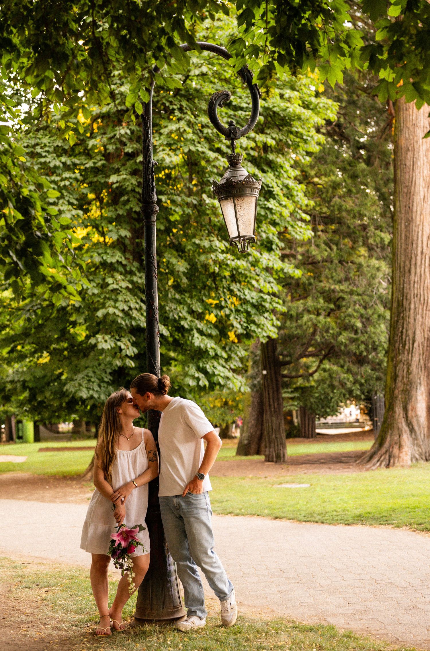 Couple sharing a kiss under a vintage park lantern surrounded by tall green trees.