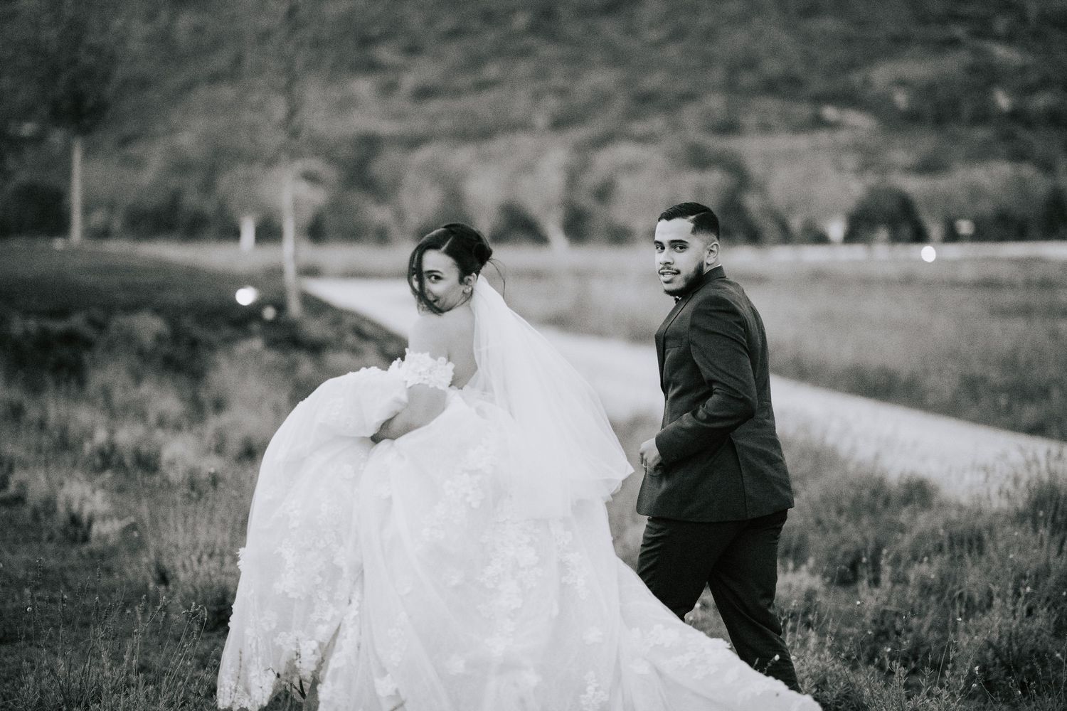 Black and white photo of bride and groom walking through a scenic outdoor field on their wedding day.