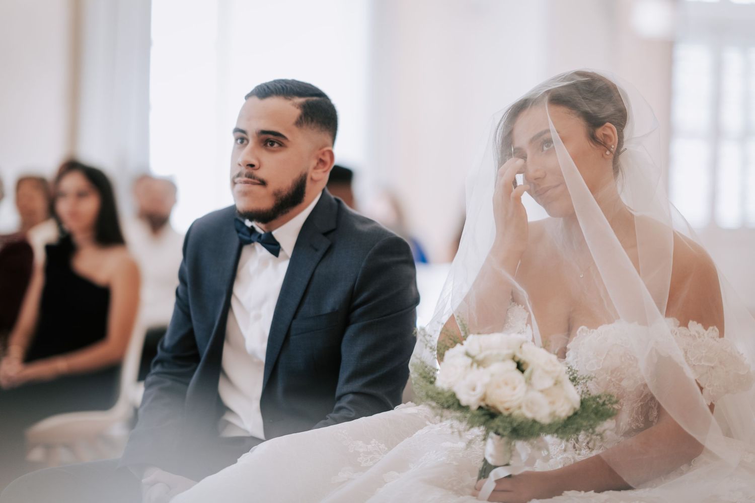 Emotional bride wipes tear while seated beside groom in navy tuxedo during wedding ceremony.