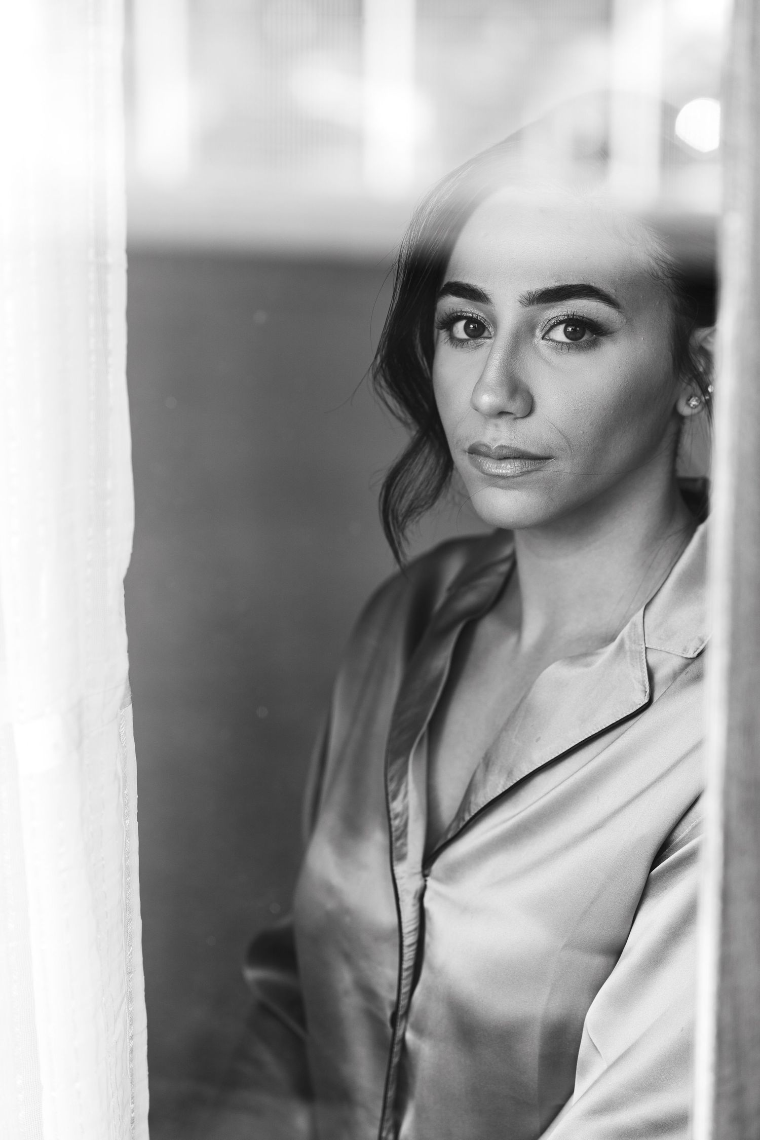 Black and white portrait of beautiful bride in satin robe gazing through window.
